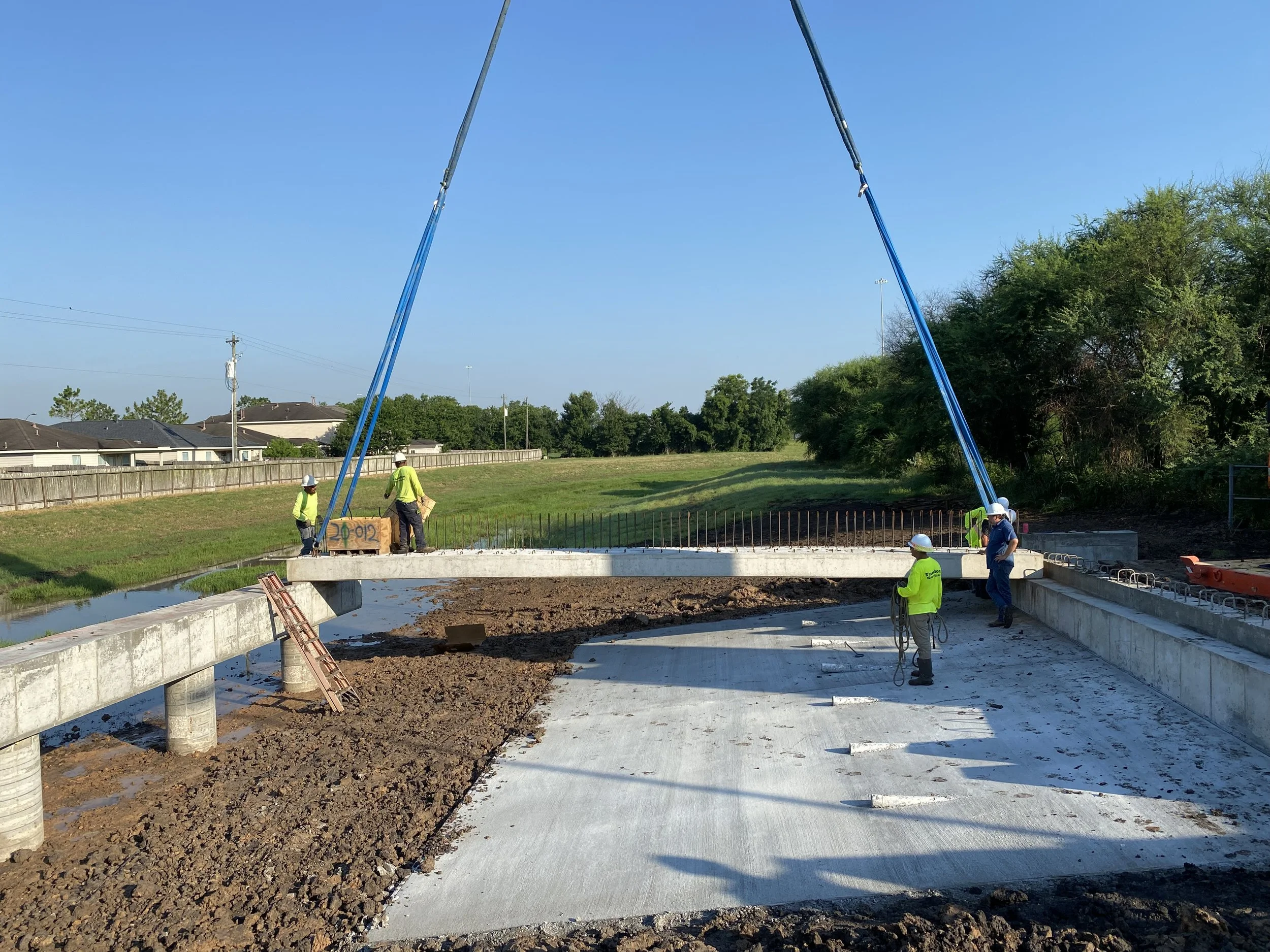 Construction workers installing a bridge over a small waterway. They are wearing safety gear and using crane equipment to position the bridge.