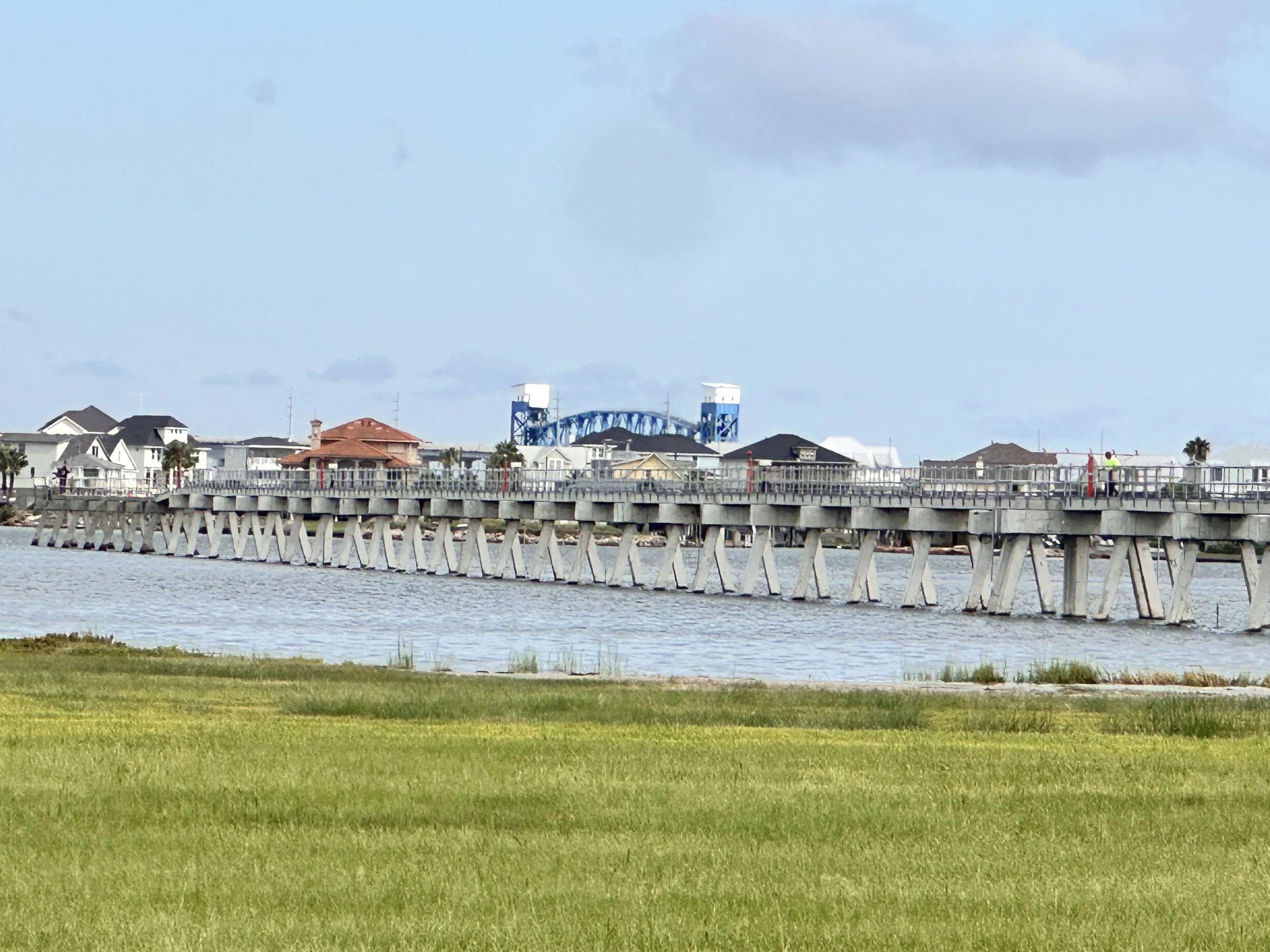 A long wooden pier extends over water with houses and structures along the shoreline, and a lagoon or water body in the foreground. Blue sky with some clouds is visible above.