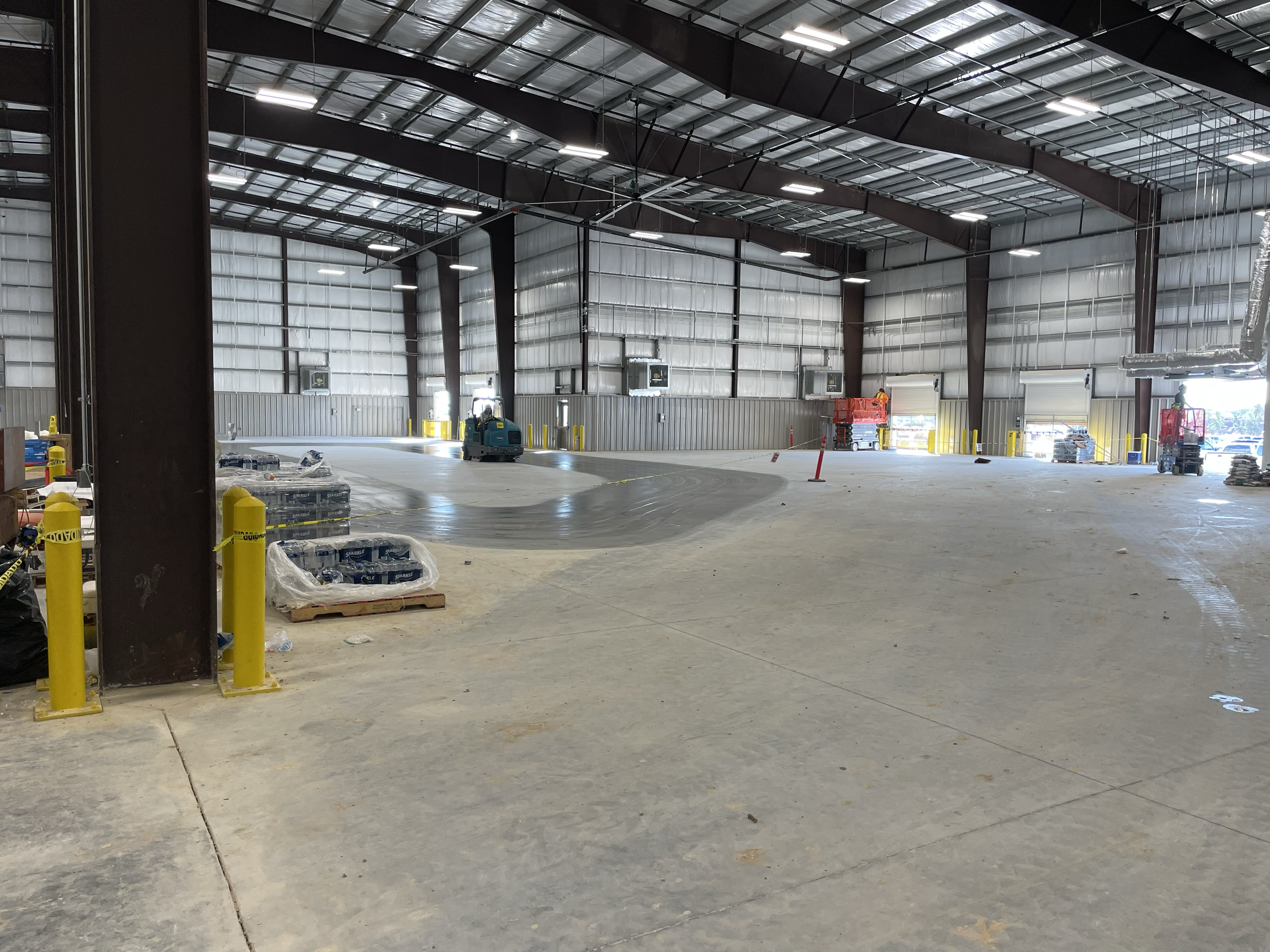 Interior of a large warehouse or industrial building under construction, with equipment, safety cones, and an autonomous floor cleaning machine.