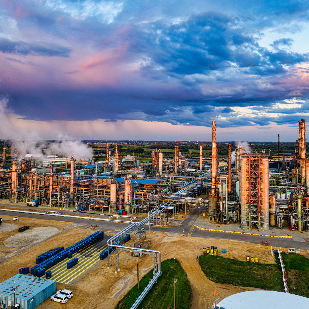 Industrial refinery with numerous pipes, smokestacks emitting smoke, and a cloudy sky overhead.