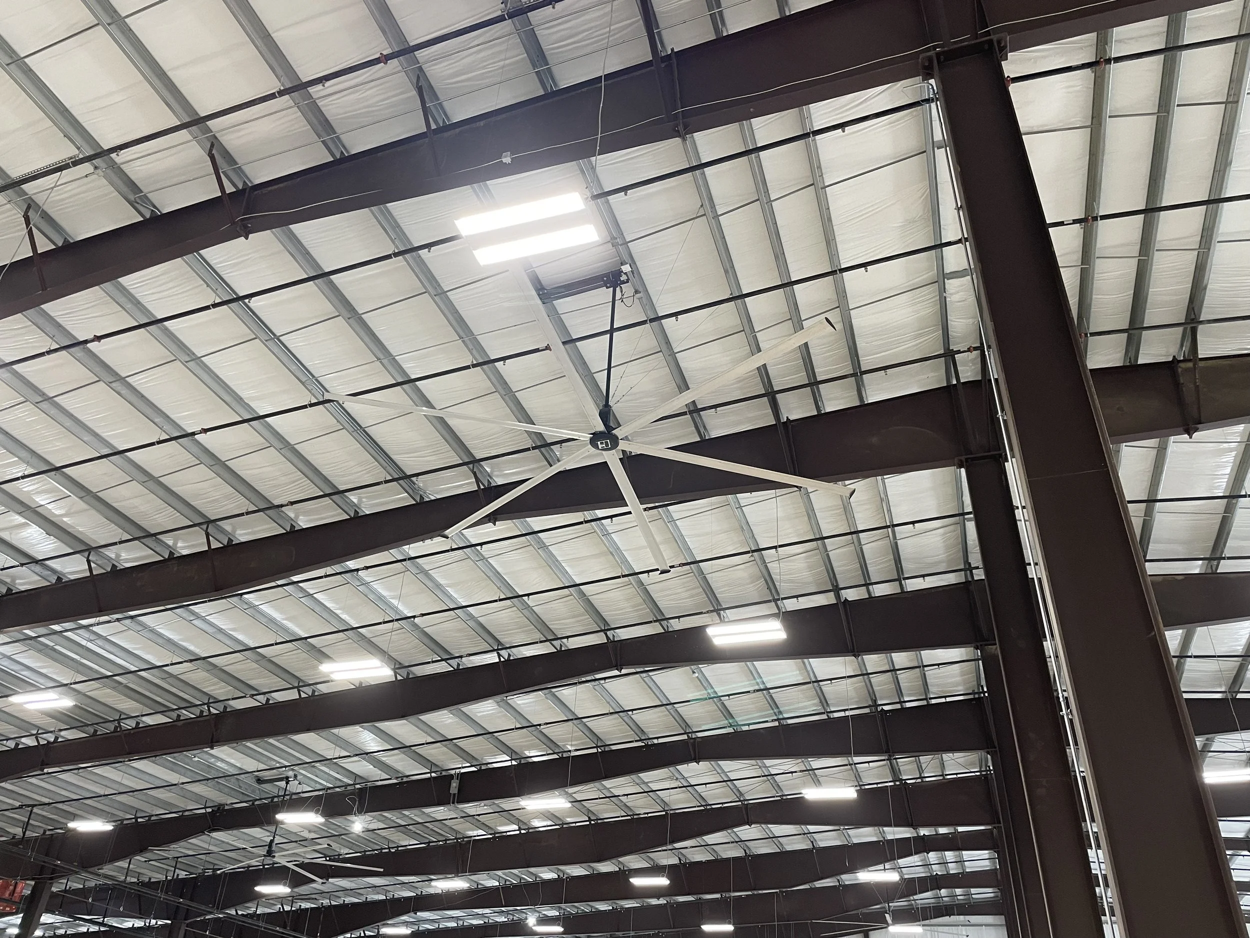 Interior of an industrial warehouse with a large ceiling fan, metal beams, and skylights or lights on the ceiling.