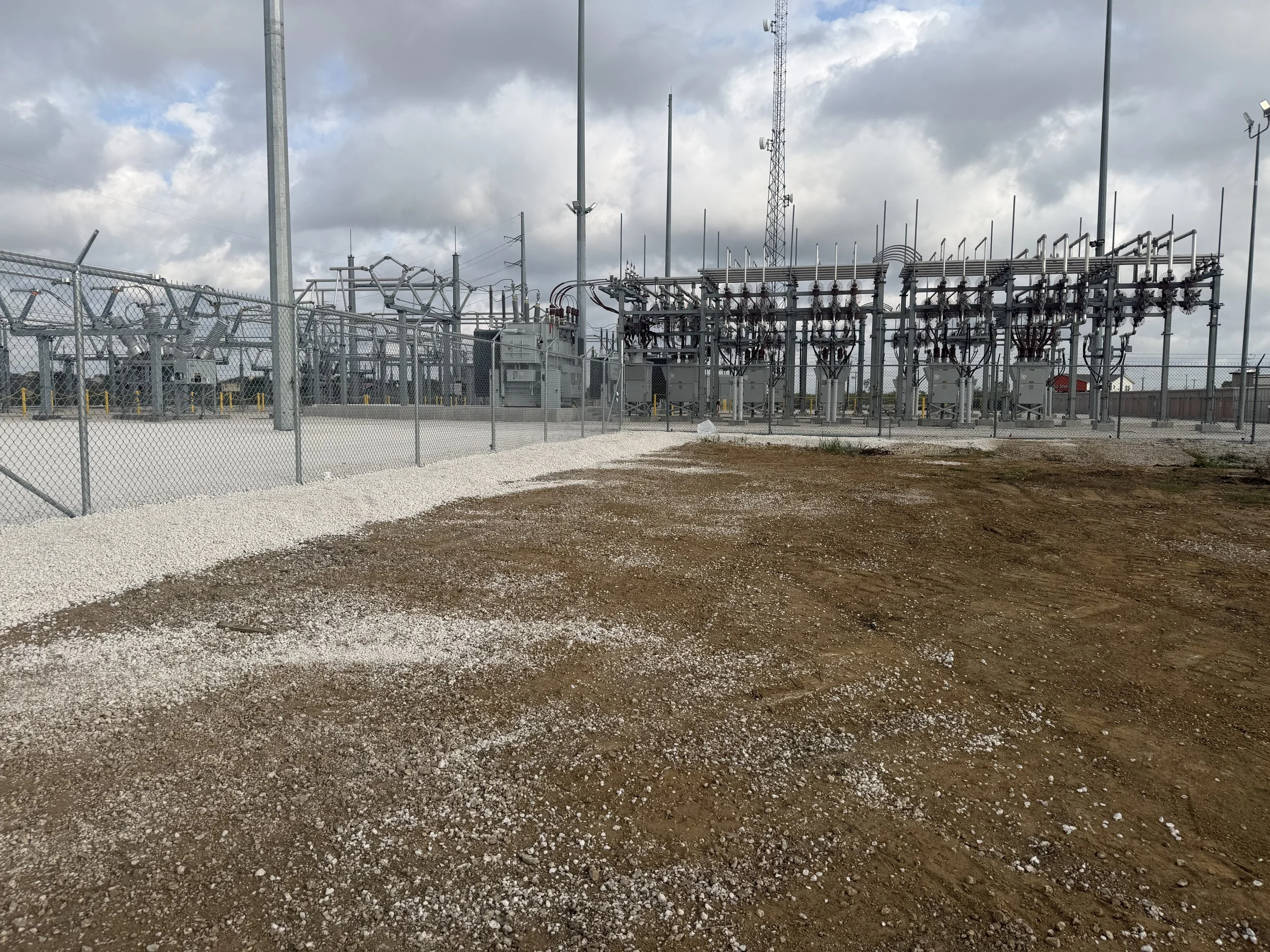 Electric substation with electrical equipment enclosed by a chain-link fence, with cloudy sky overhead.