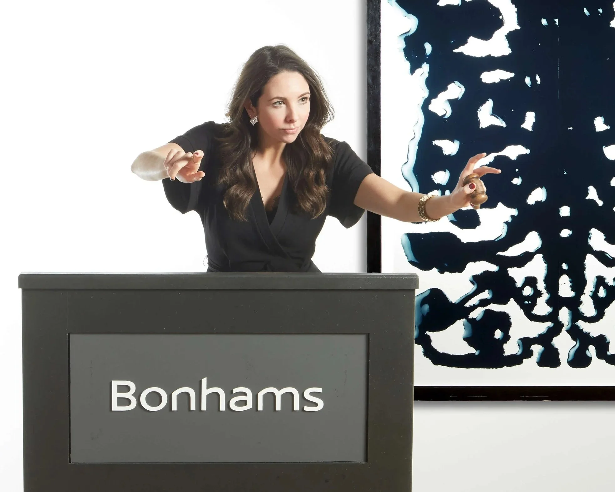 Jacqueline at an art auction, standing behind a podium with 'Bonhams' written on it, pointing toward a piece of modern abstract art in the background.