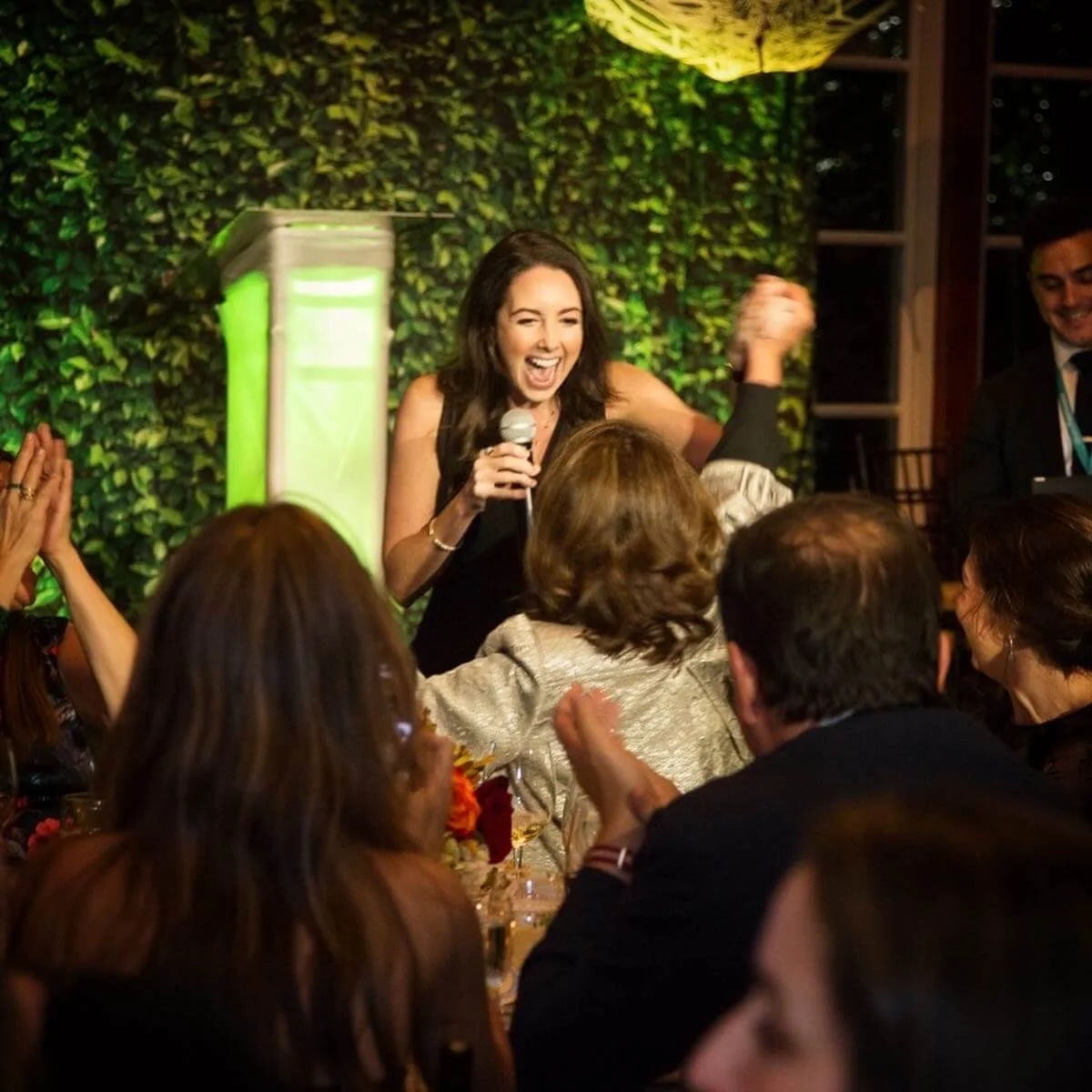 Jacqueline holding a microphone, smiling, standing in front of a green leafy backdrop, surrounded by a group of people at a celebration or event.