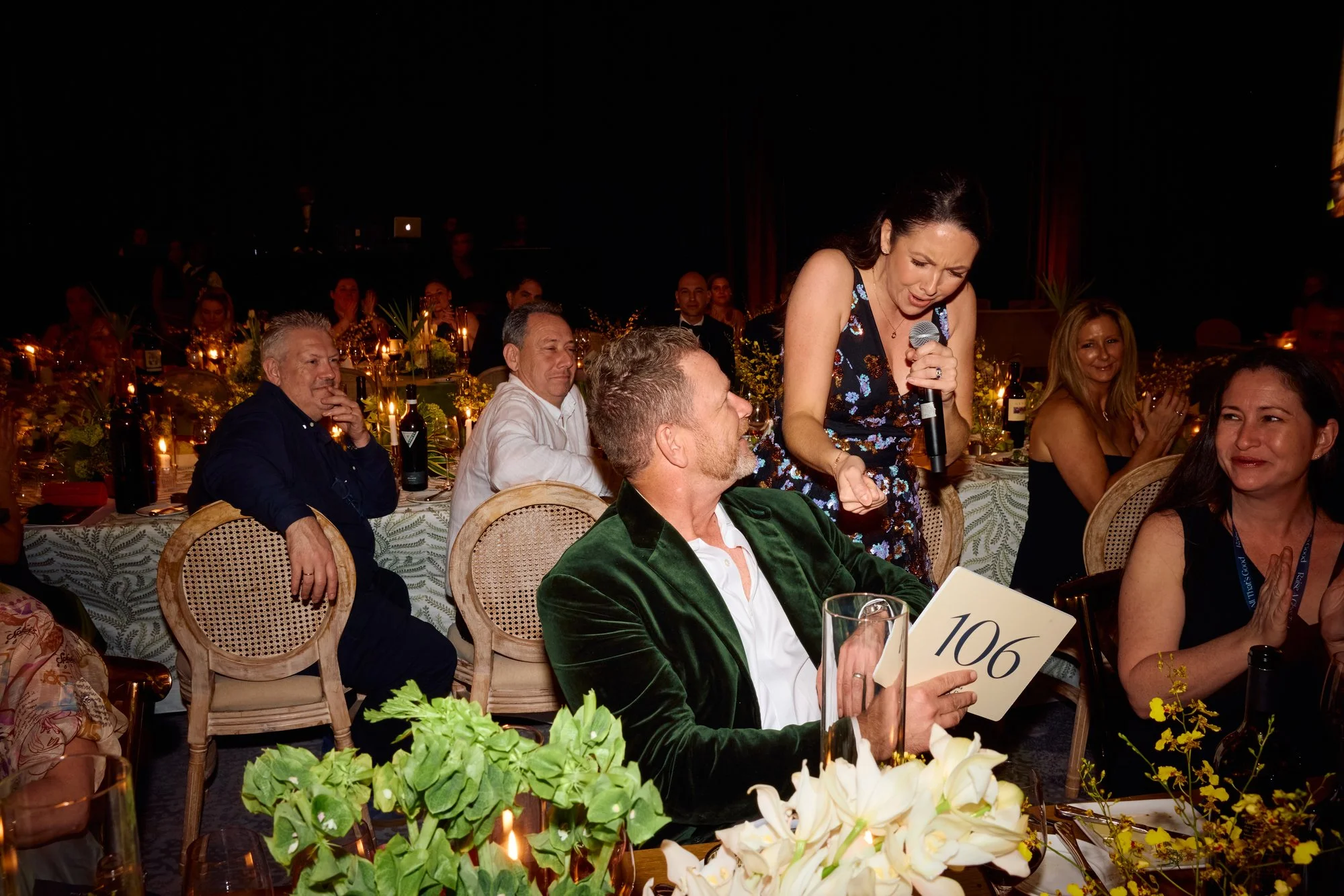 Jacqueline holding a microphone leans in towards a man seated at a table with a sign labeled '106' at a formal event, with other guests sitting and clapping around them, decorated with flowers and candles.