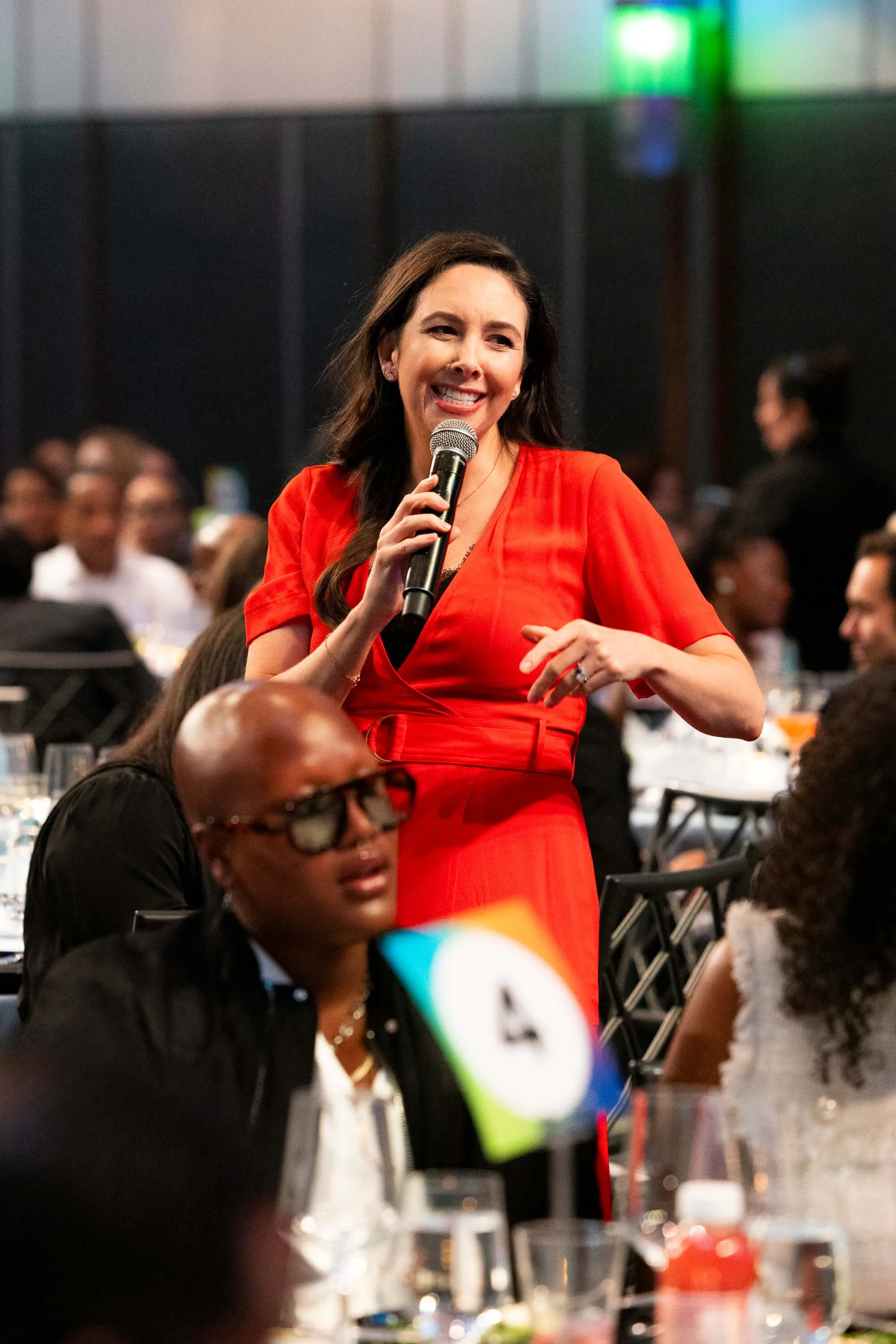 Jacqueline in a red dress holding a microphone and speaking at a banquet or dinner event with tables, glasses, and people in the background.