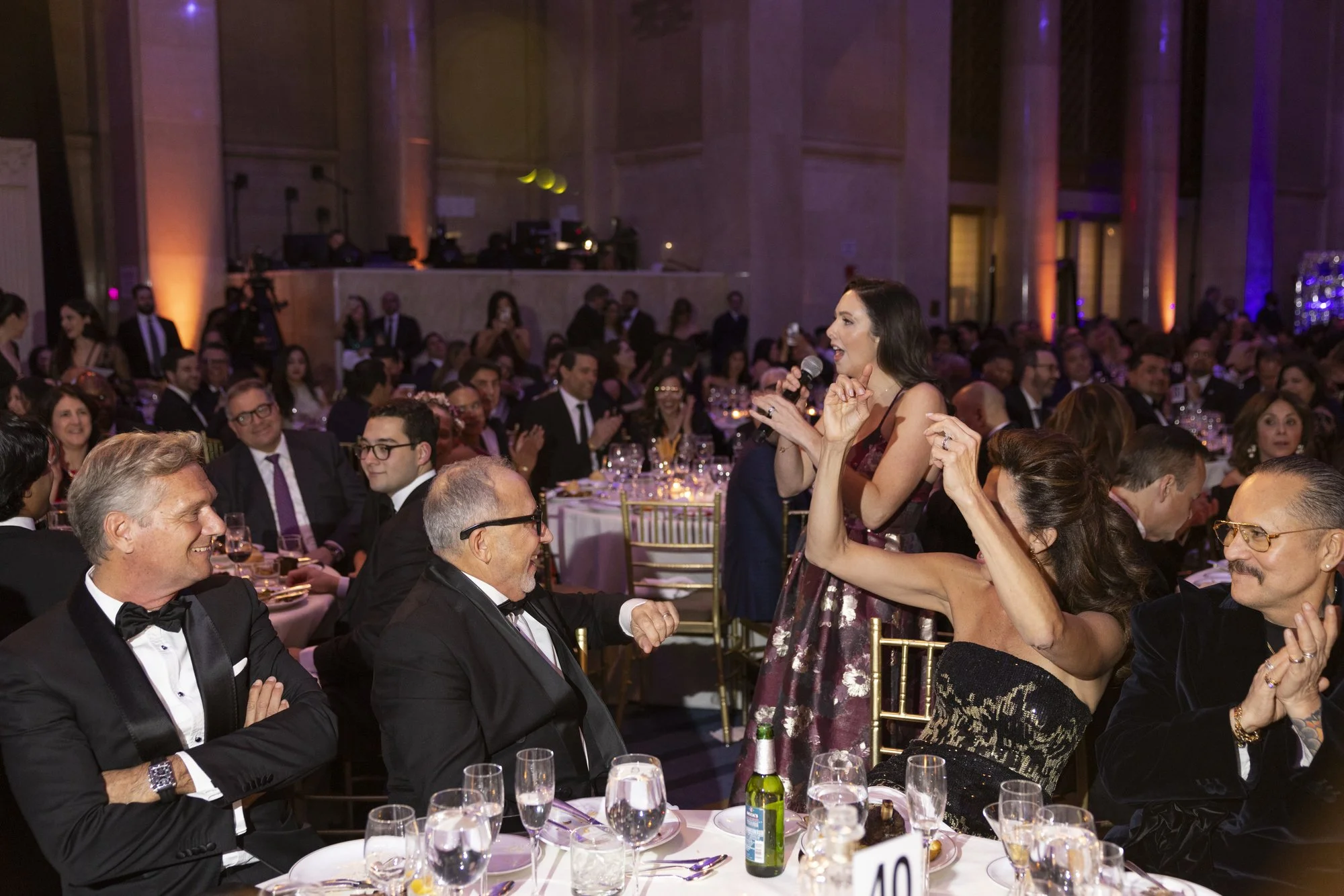 Jacqueline speaking into a microphone at a formal event, surrounded by seated guests in tuxedos and evening gowns, with a large crowd in a banquet hall with purple and orange lighting.