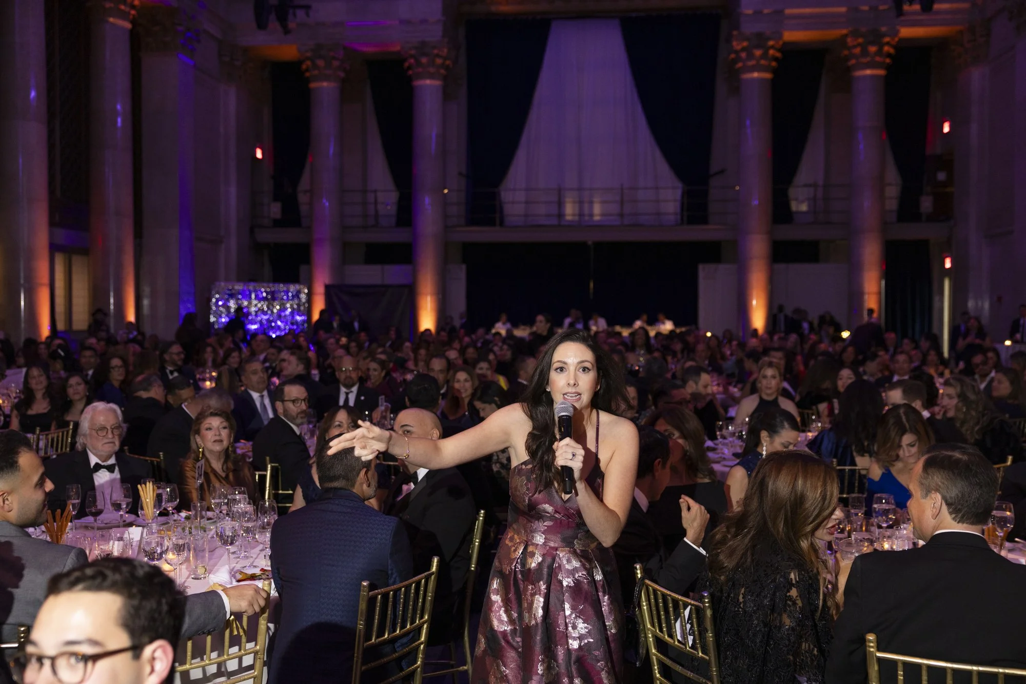 Jacqueline speaking into microphone at formal event with many seated guests in elegant attire, large decorated hall with purple and warm lighting.