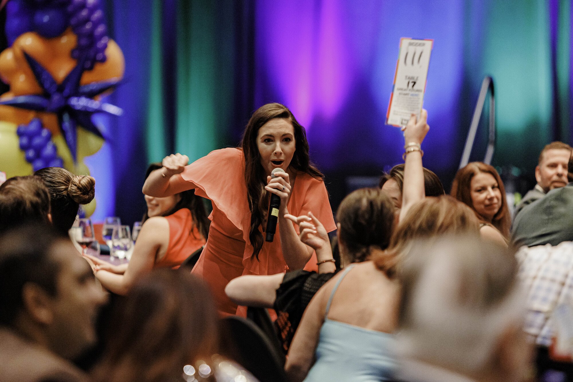 Jacqueline holding a microphone and raising her hand while speaking at a lively event. She is surrounded by seated attendees, one of whom is raising a bidding paddle. The background features colorful decorations and a vividly lit stage.