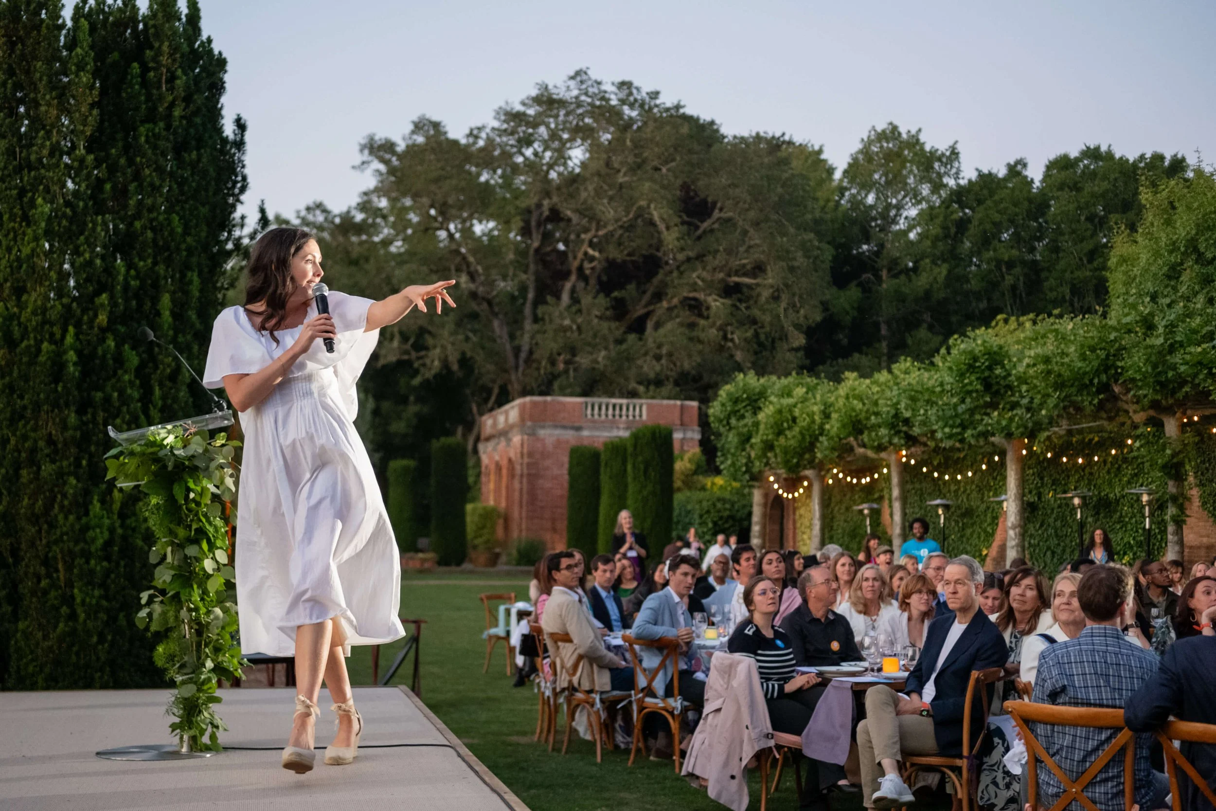 Jacqueline in a white dress speaking into a microphone on an outdoor stage, with an audience sitting at tables and a garden with string lights in the background.