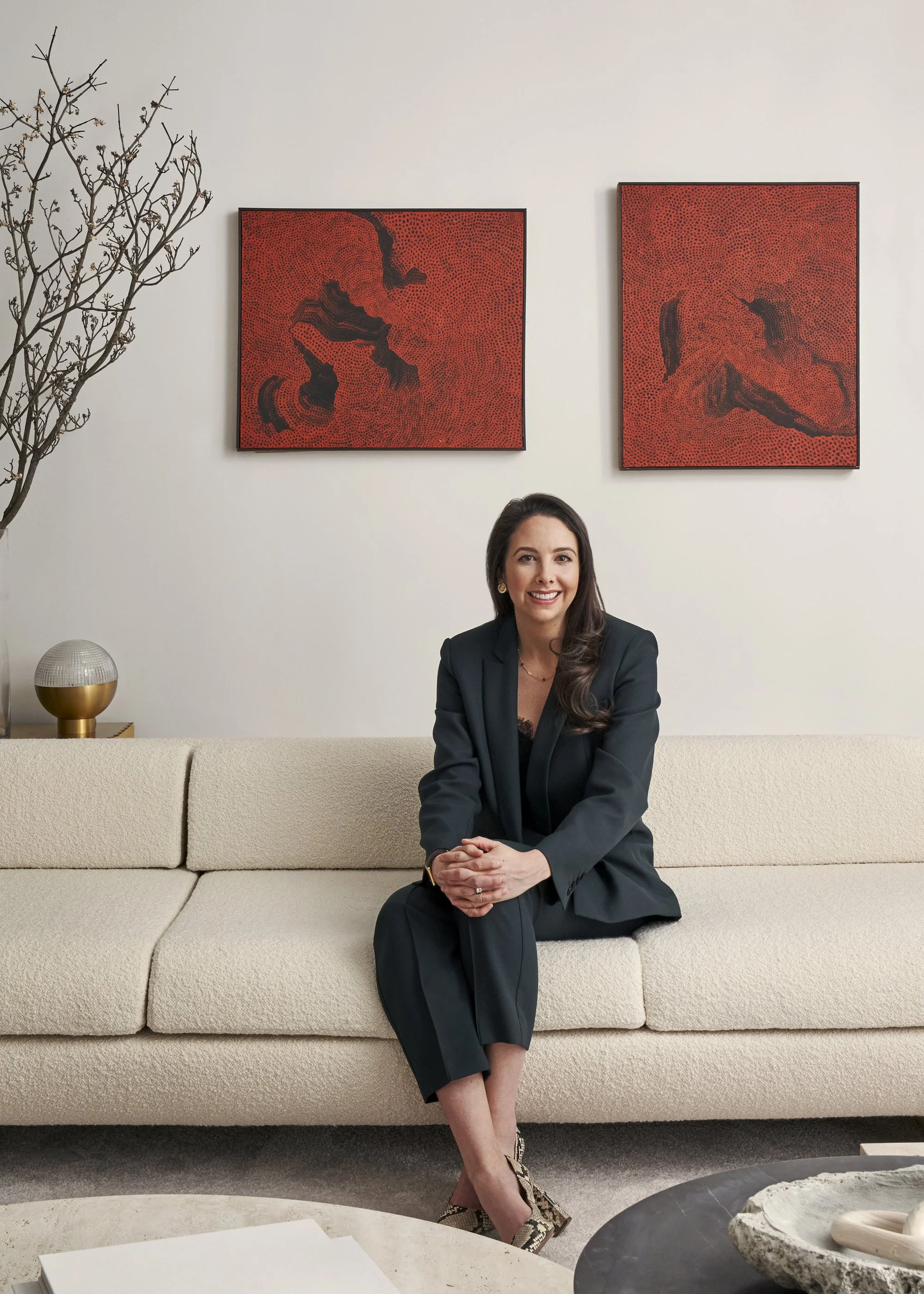 Jacqueline Towers-Perkins sitting on a beige sofa in a modern living room, smiling at the camera. She is dressed in a black suit and patterned high heels with dark red art in the background.