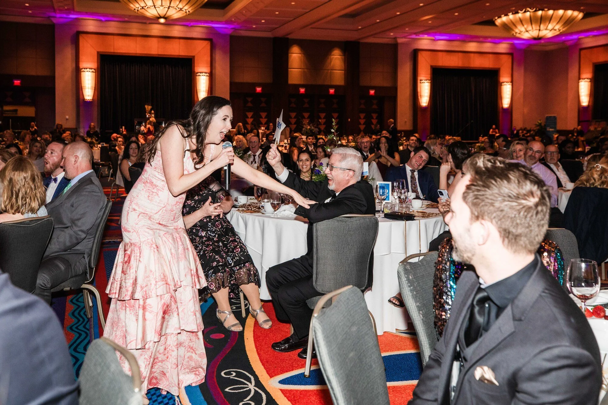 Jacqueline with a microphone while reaching out to a seated man in a tuxedo at a formal dinner event. The background shows many other attendees seated at tables, clapping and smiling in a decorated banquet hall with warm lighting.