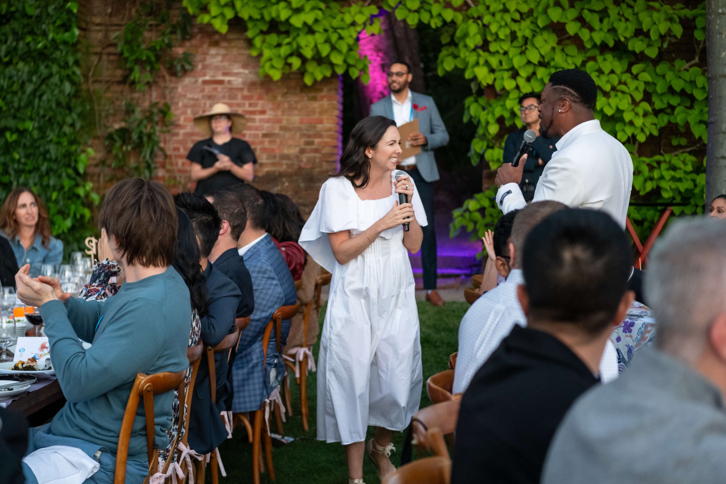 A wedding reception outside with guests seated at long tables, Jacqueline in a white dress holding a microphone and smiling, and a man in a white suit speaking into a microphone, with a green leafy background and brick wall.