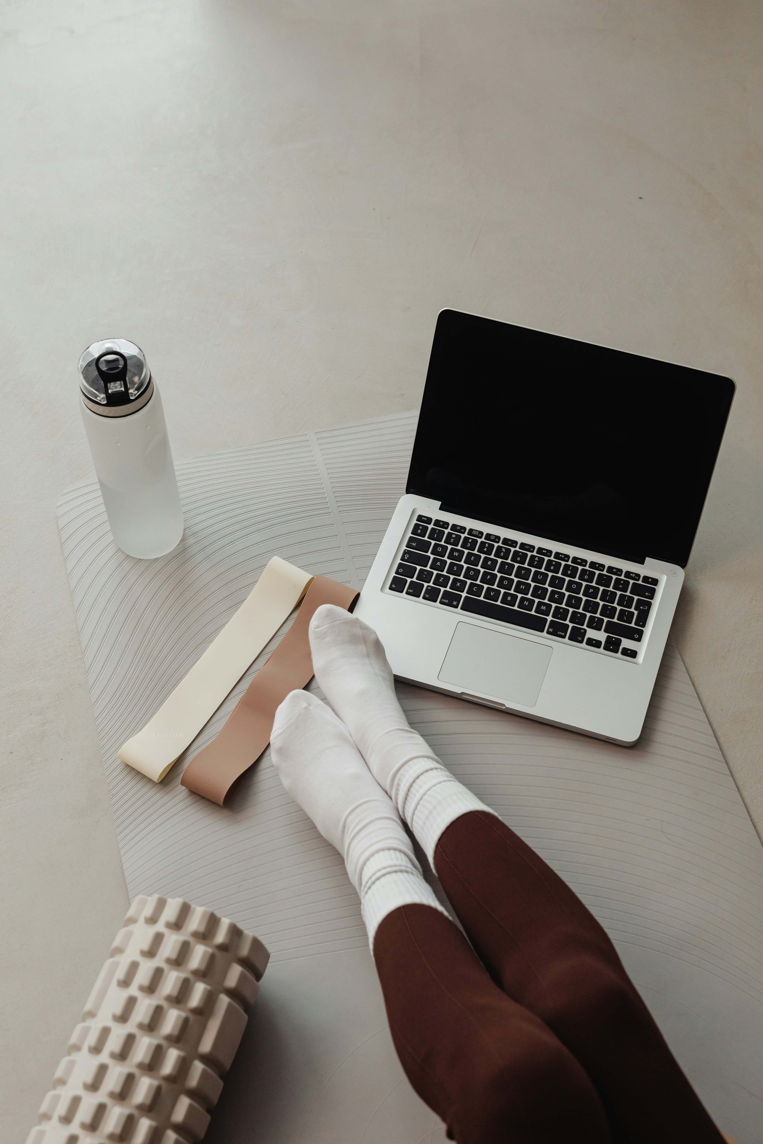 Overhead view of a workspace with a person's legs in white socks and maroon pants, a silver laptop, a white water bottle, two fabric swatches, and a foam roller on a textured light grey surface.