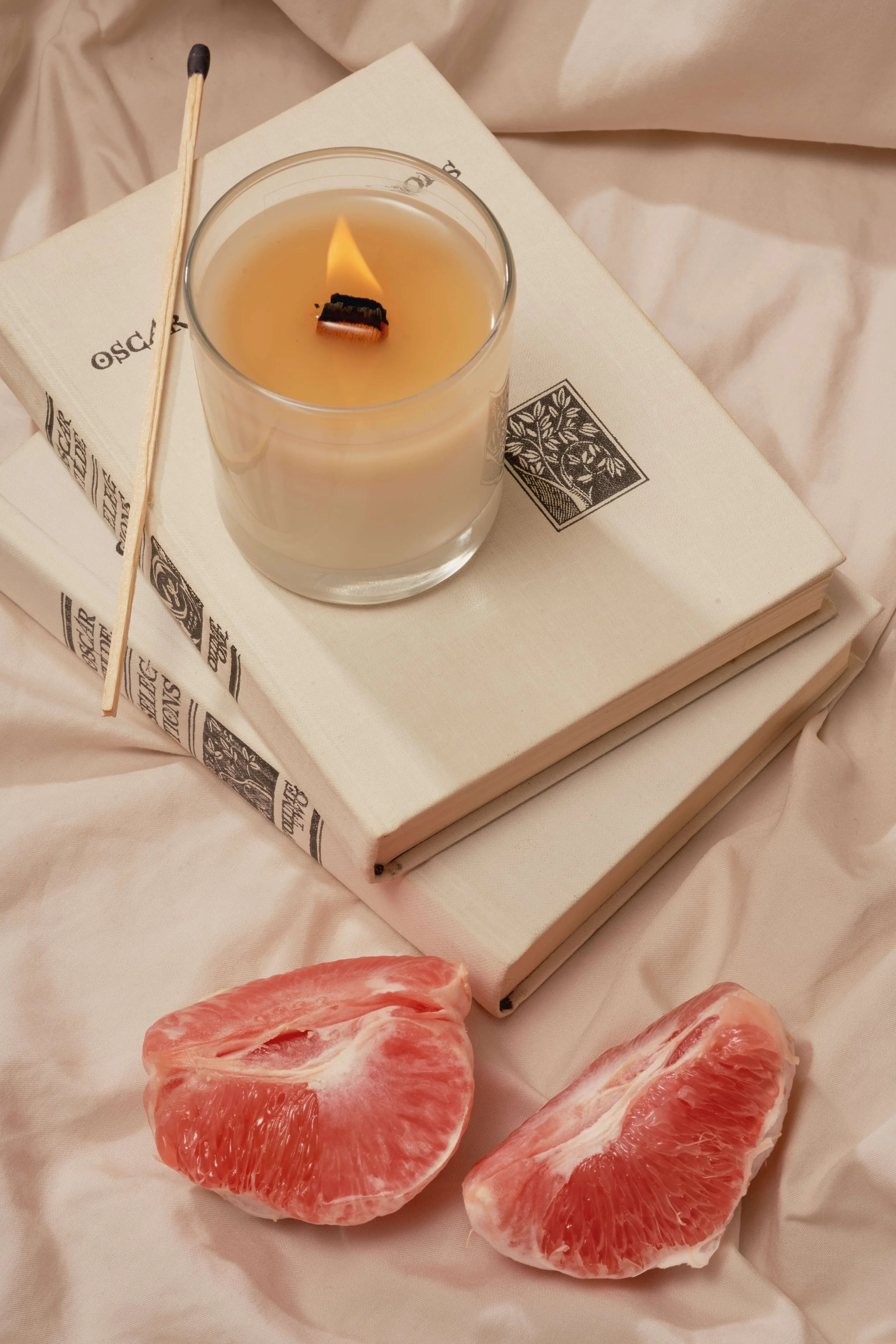 A lit candle in a glass container placed on top of a stack of two books with white covers, one titled 'OSCAR' and featuring a black tree illustration, on a pink fabric surface. Two halves of a pink grapefruit are placed in front of the books.