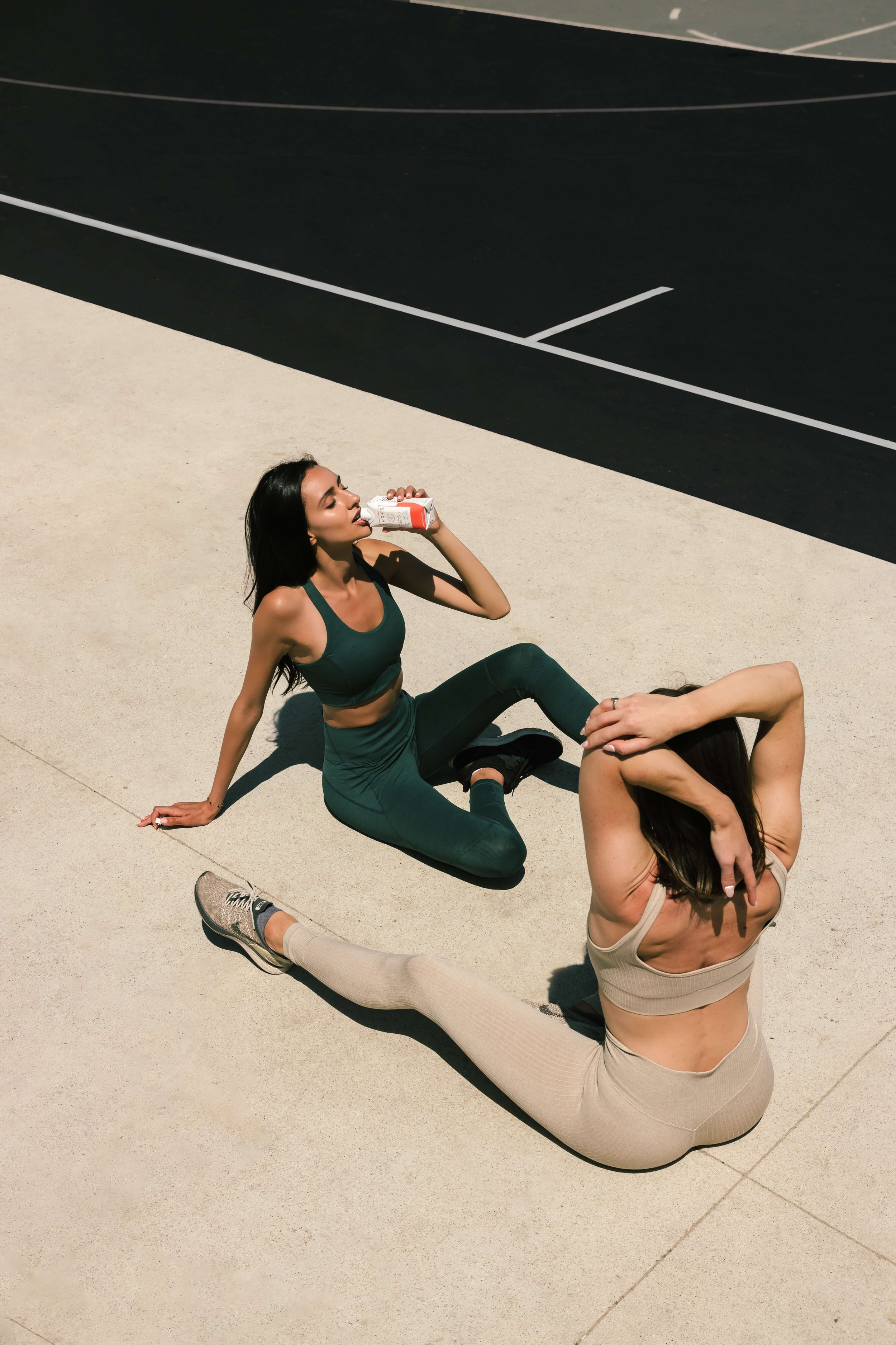 Two women in workout clothes sitting on a concrete sidewalk near a black asphalt parking lot. One woman is drinking from a white and red bottle, and the other woman is stretching with her hands over her head.