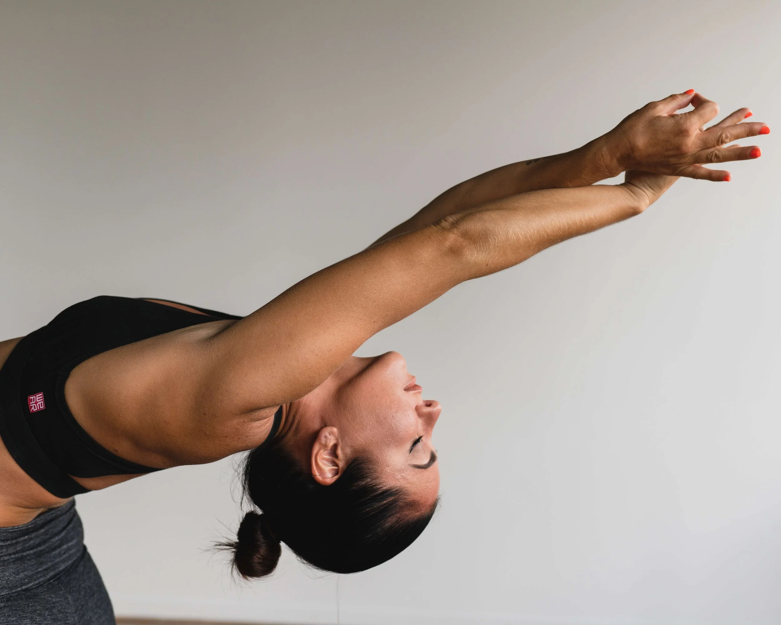 A woman doing a forward bend yoga pose, reaching her arms overhead with hands clasped, wearing a black sports bra and gray leggings.
