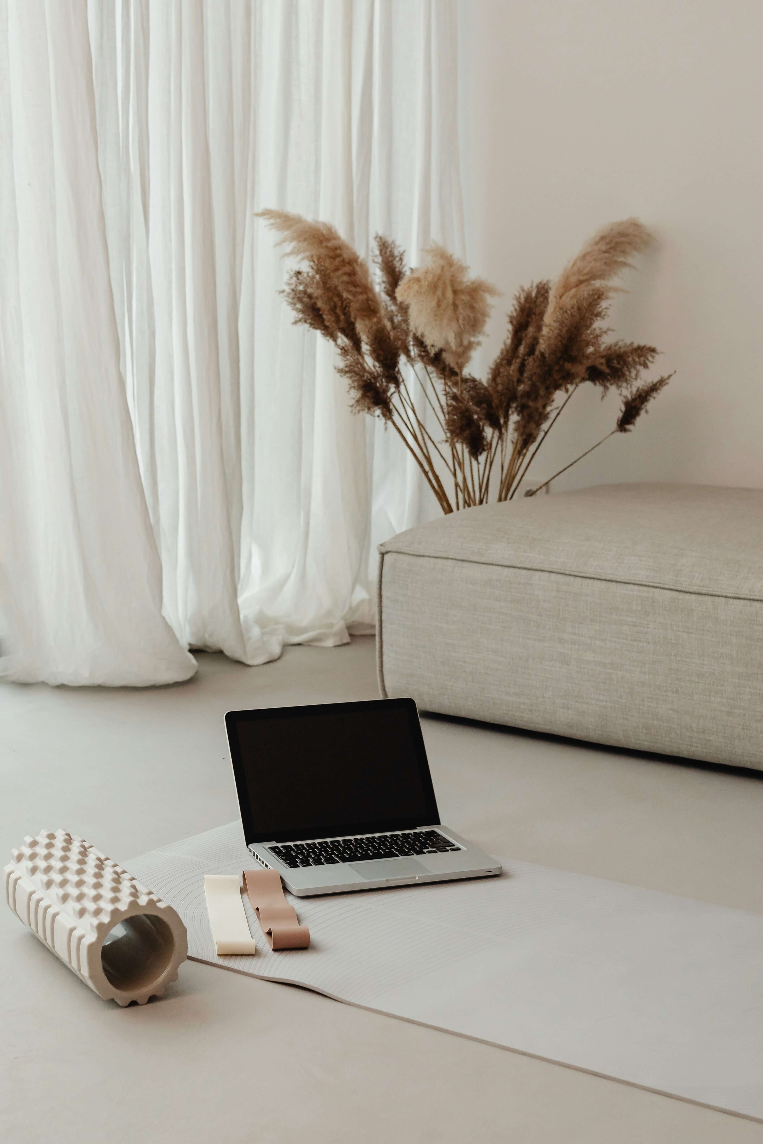 Living room with a MacBook on a white yoga mat, a foam roller, and resistance bands, with a beige sofa and dried pampas grass in the background.