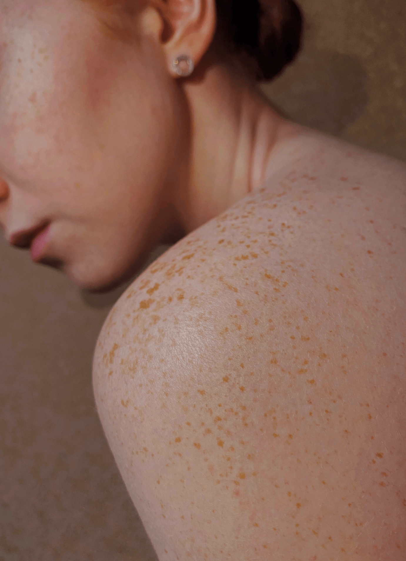 Close-up of a woman's shoulder and face, showing natural freckles and a small earring, with her head turned downward.