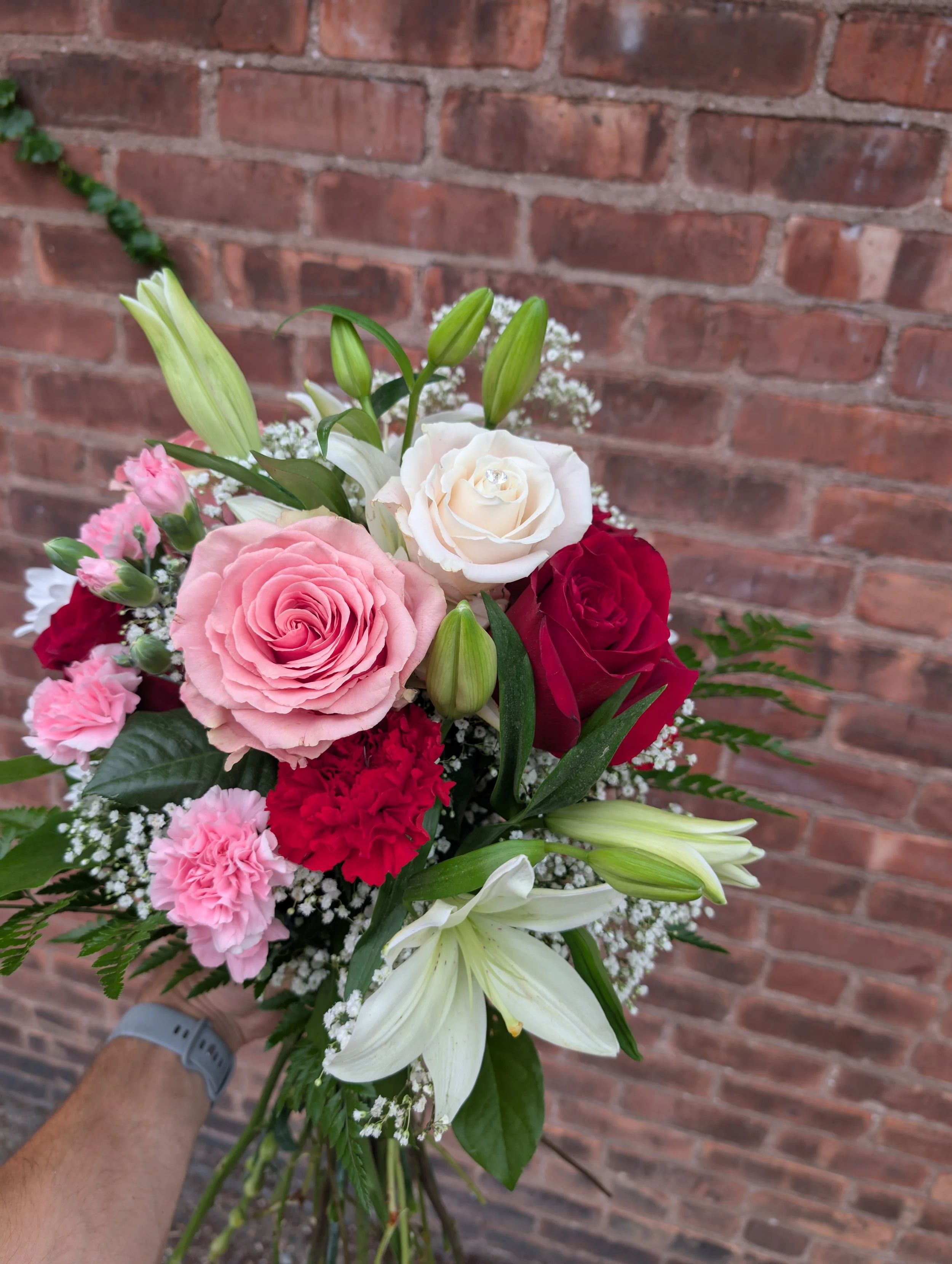 A bouquet of mixed flowers including pink roses, white lilies, white roses, red roses, pink carnations, baby's breath, and assorted green foliage.