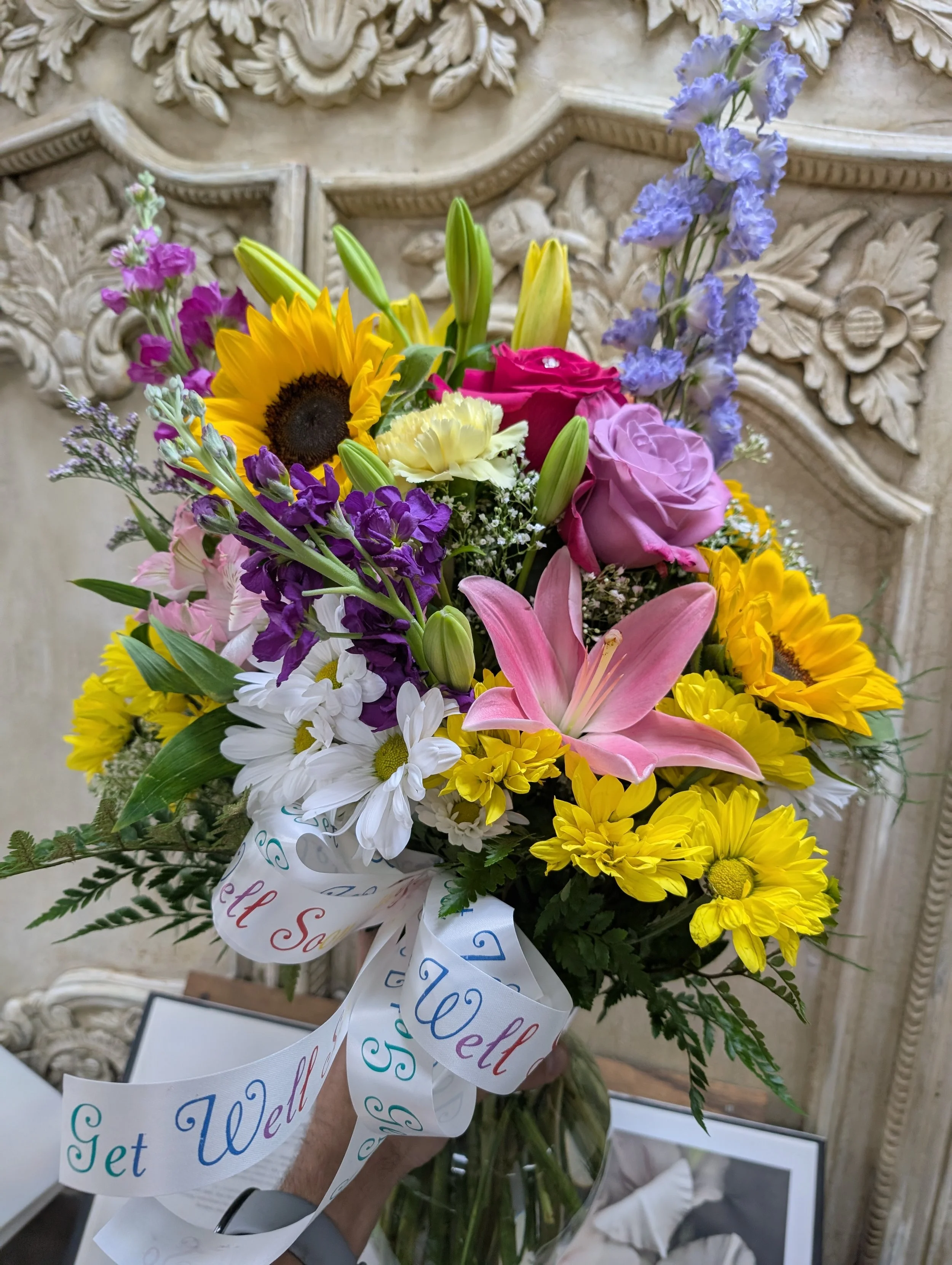 A vibrant bouquet of mixed flowers including sunflowers, pink lilies, roses, daisies, snapdragons, and other colorful blooms, wrapped with a white ribbon that says 'Get Well.'