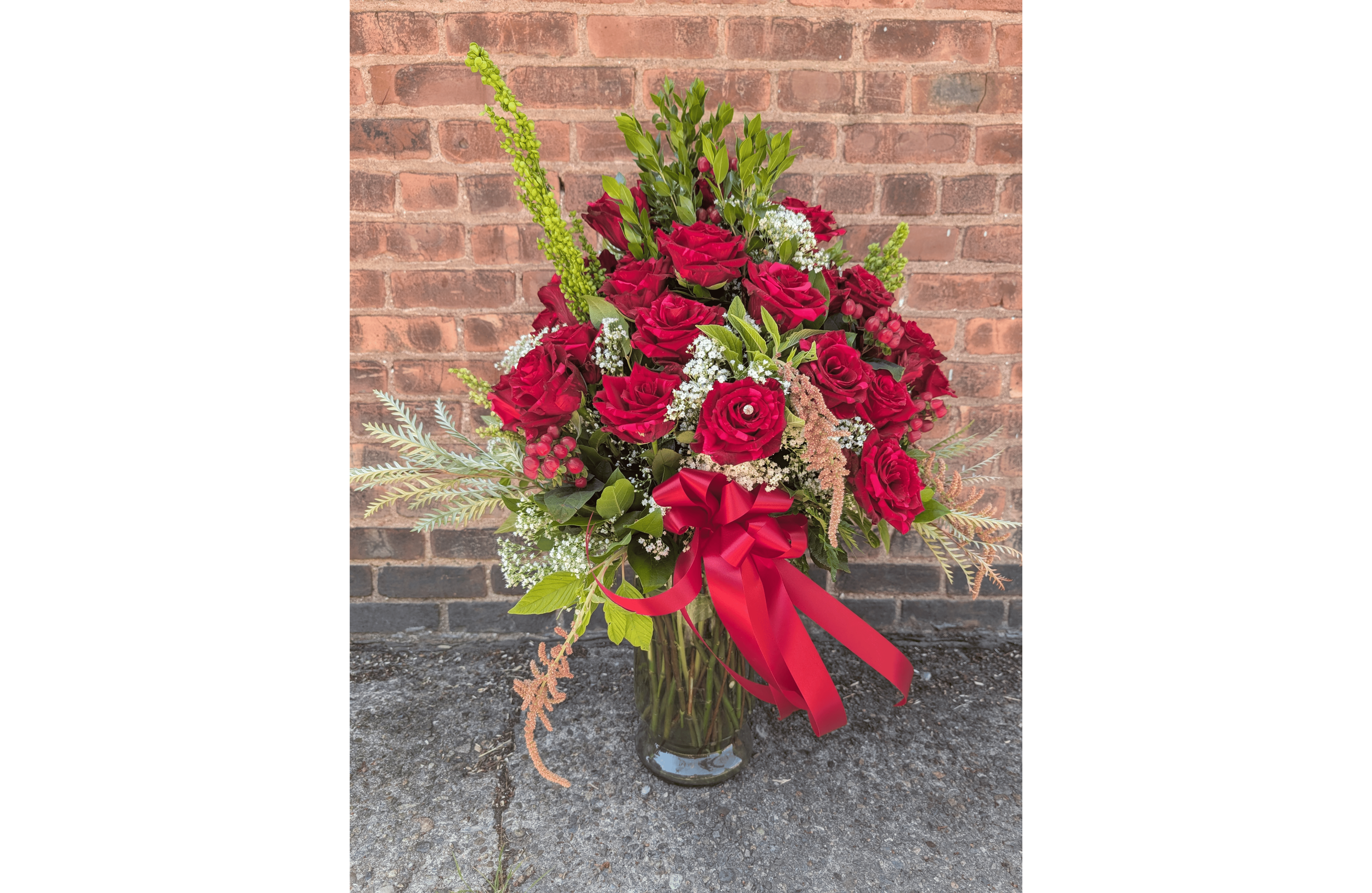 A bouquet of red roses, white filler flowers, and greenery arranged in a glass vase, adorned with a red ribbon bow.