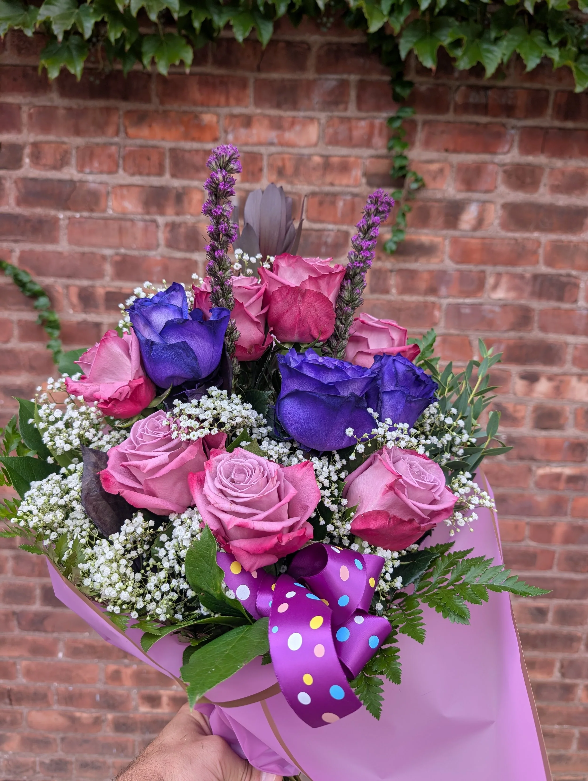 A colorful bouquet of pink roses, purple lisianthus, lavender, baby's breath, and green foliage, wrapped in pink paper with a purple polka-dotted ribbon.