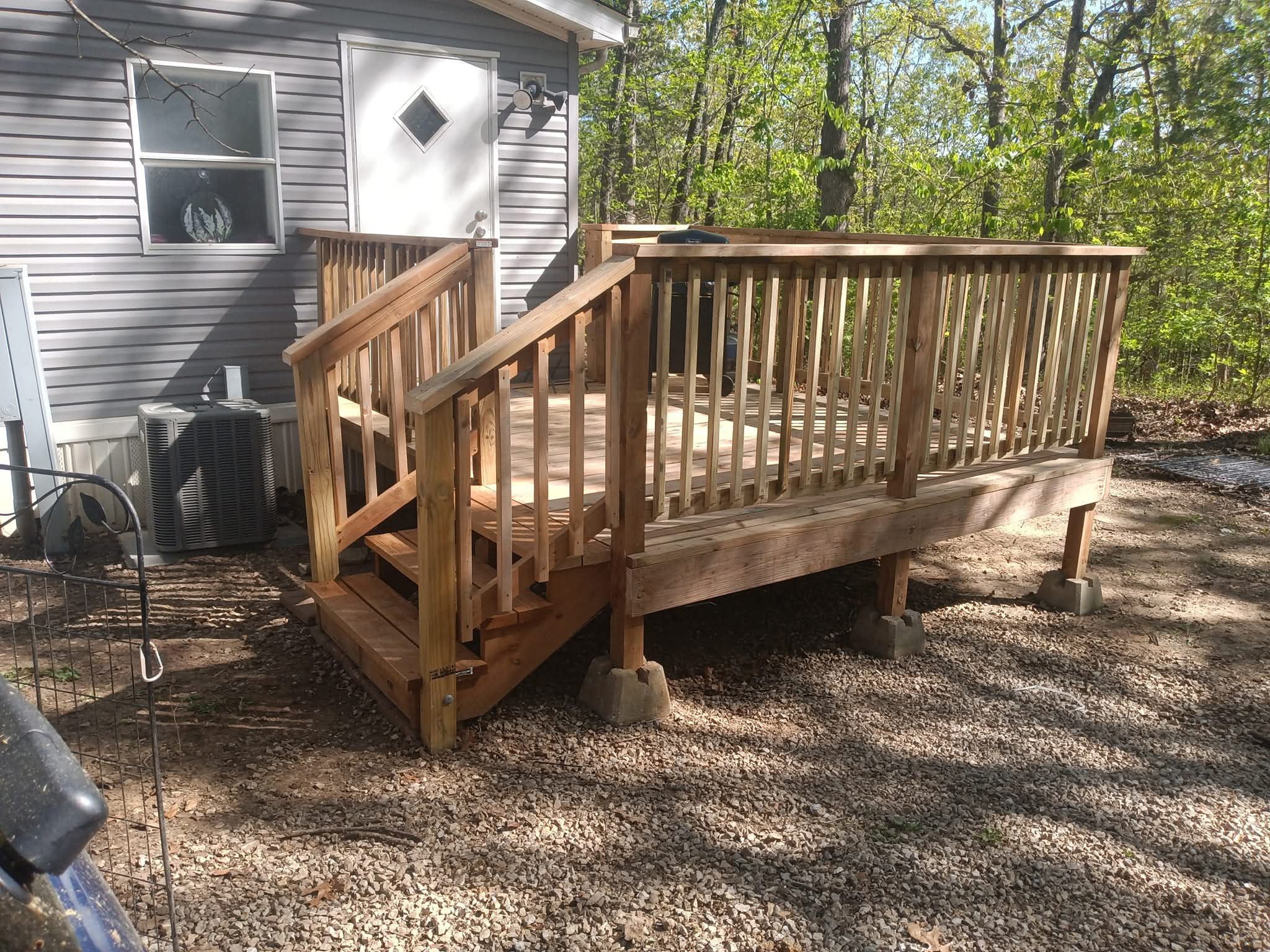 Wooden deck with stairs attached to the back of a house, surrounded by trees and dirt ground.
