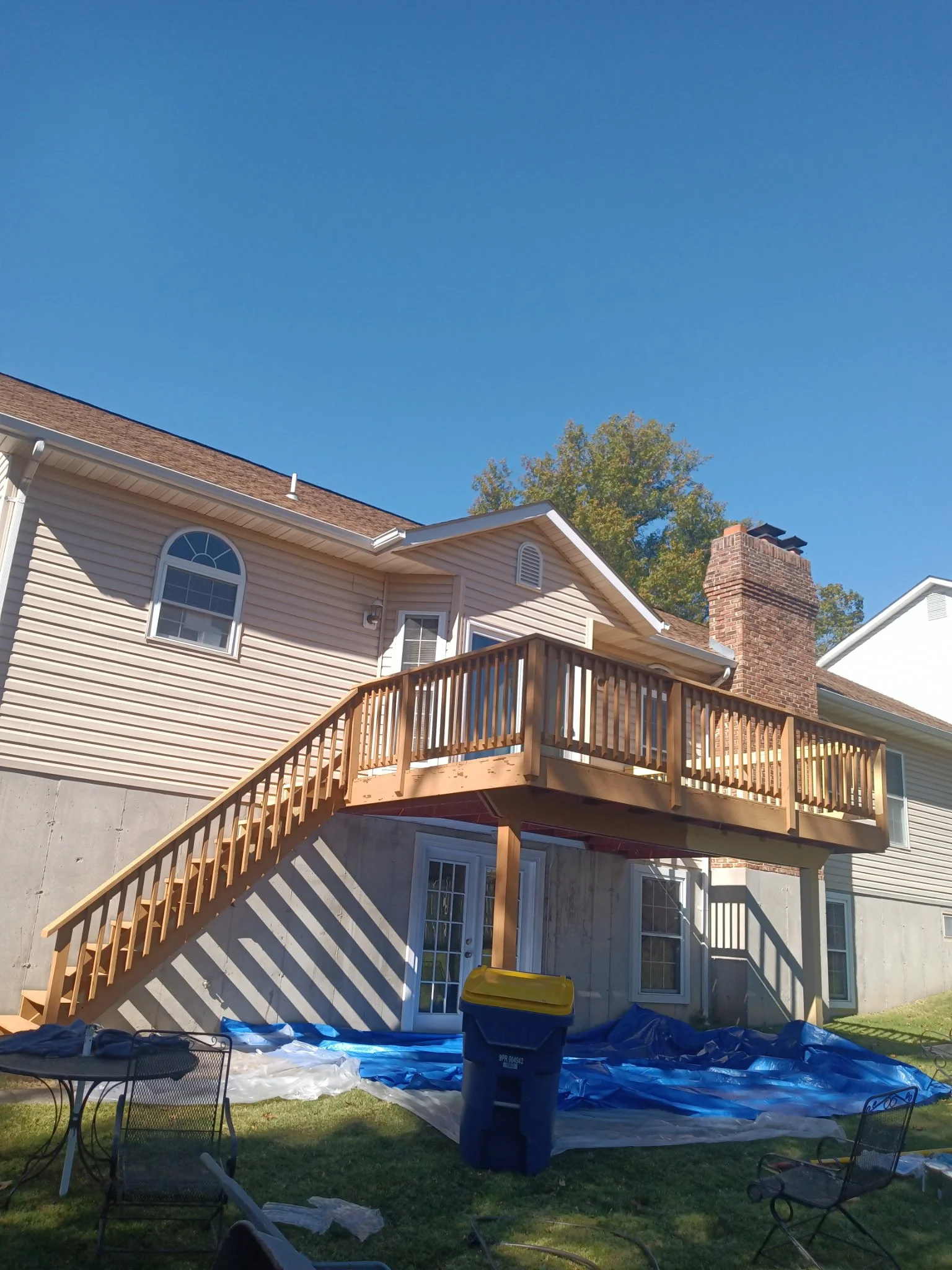 Back of a house with a new wooden deck and stairs, a brick chimney, and a blue tarp on the ground, with outdoor chairs and a trash bin under a clear blue sky.
