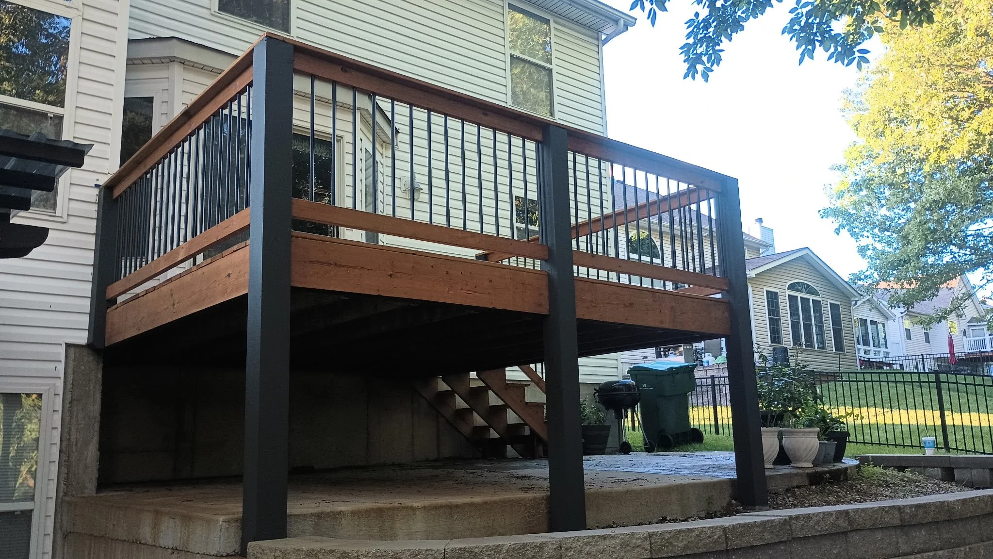 Backyard wooden deck with black metal railing and stairs, surrounded by green lawn and neighboring houses, under a tree with blue sky.