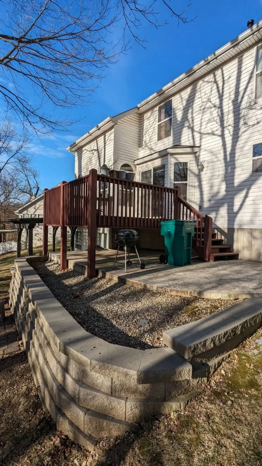 Backyard with a wooden deck, grill, green trash bin, and stone retaining wall on a sunny day with leafless trees casting shadows on the house.