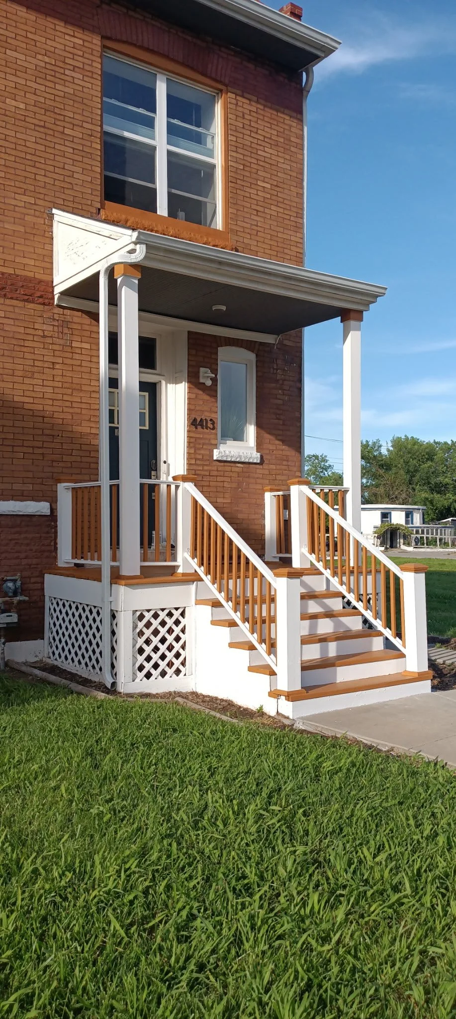 Front porch of a brick house with white and wood-colored railing and stairs leading to a black door, glass window, and a small overhang, on a sunny day.