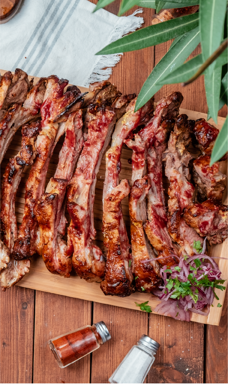 Costelas assadas Carvalho dispostas sobre uma tábua de madeira, decoradas com cebola roxa, coentro e pimenta em vidro ao lado, sobre mesa de madeira.