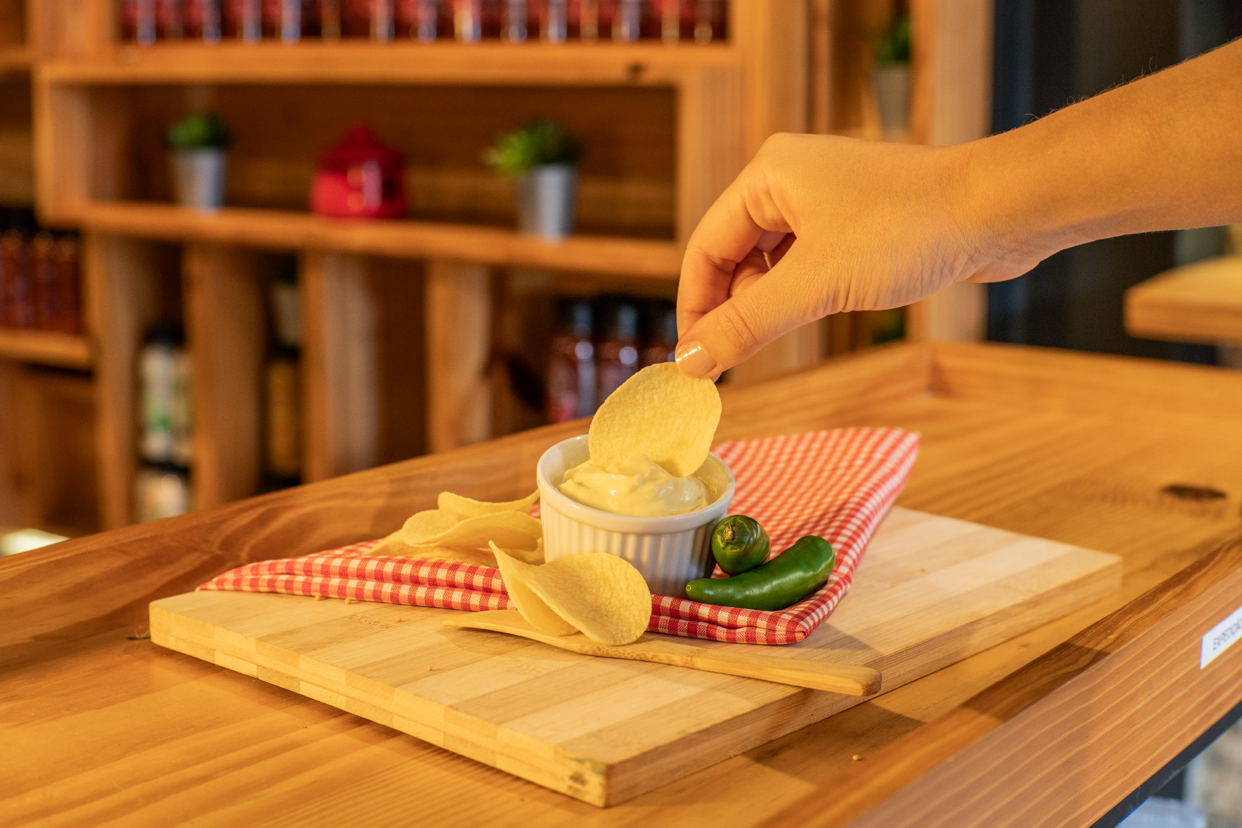Mão colocando uma batata chip em um potinho de maionese. Ao lado, há uma pimenta verde e um mini pimentão em uma bandeja de madeira, com uma toalha xadrez vermelha e branca.