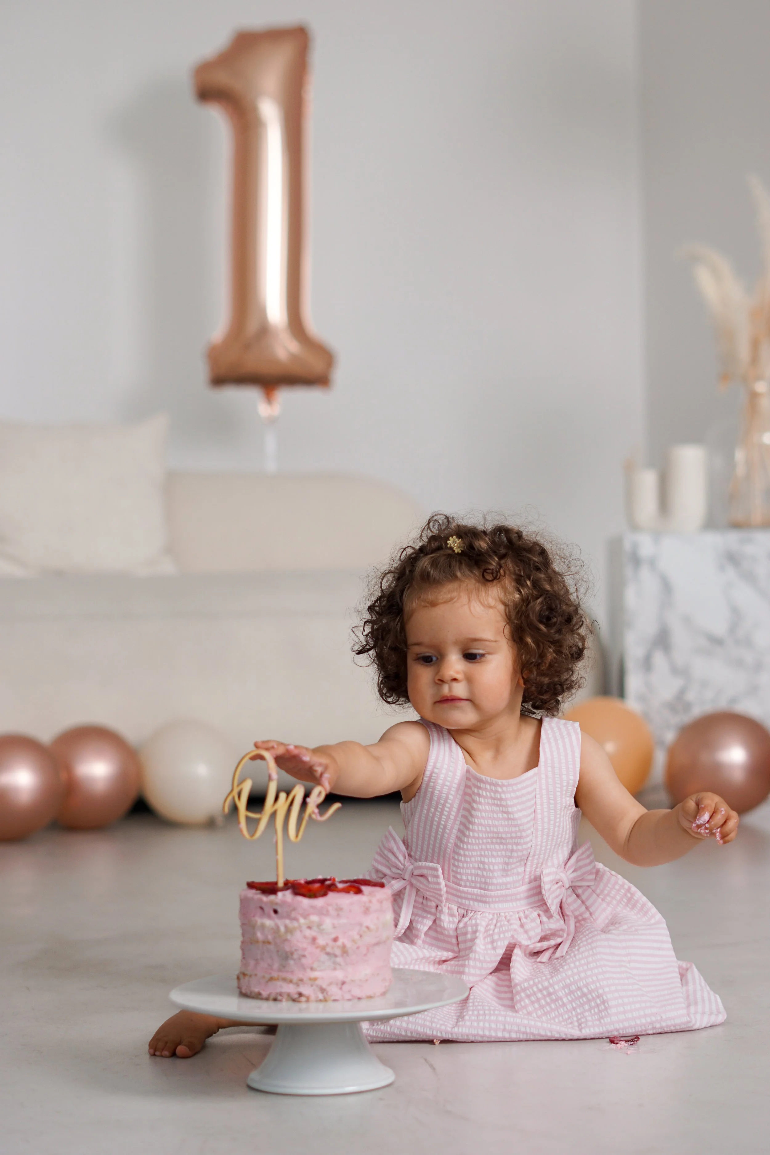 A young girl with curly hair in a pink dress sits on the floor, reaching to touch a pink birthday cake with a topper that says 'one.' There are balloons in the background, including a large number '1' balloon.