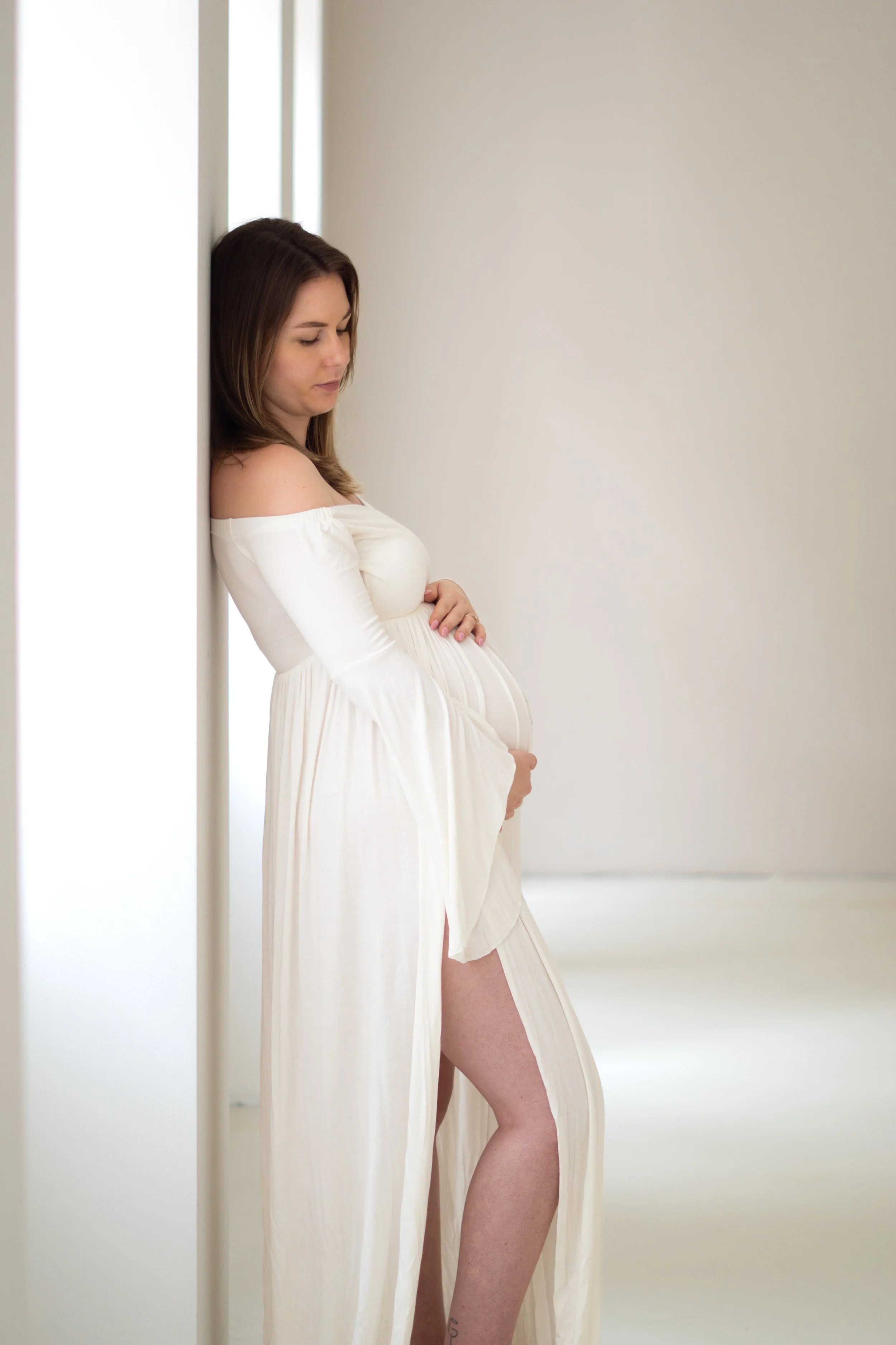 Pregnant woman in a white dress placing her hand on her belly while leaning against a wall in a brightly lit room.