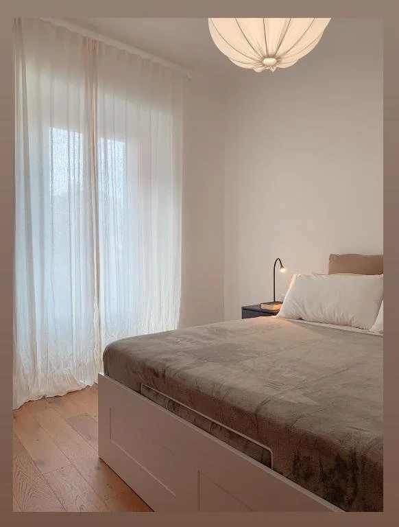 Camera da letto minimalista con letto matrimoniale, cuscino e coperta beige, lampada da tavolo accesa, finestra con tendaggi trasparenti, soffitto con lampada di carta.