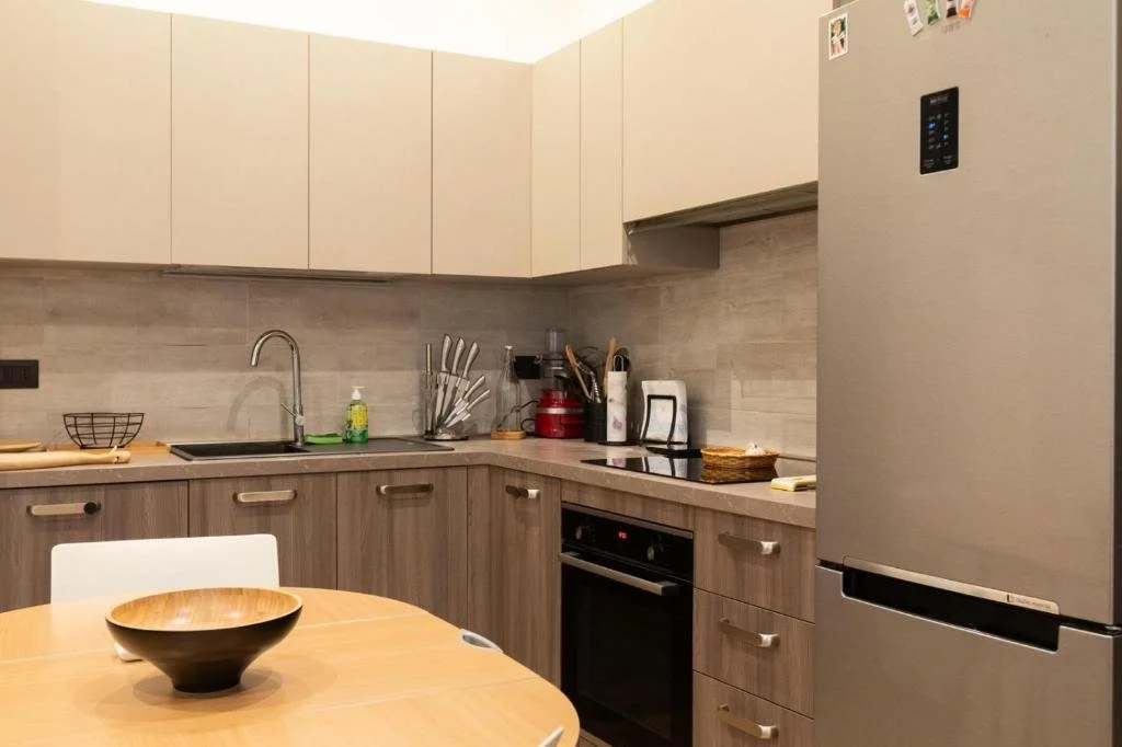 Cucina moderna con ripiani in legno chiaro, lavello, frigorifero, vari utensili e piccoli elettrodomestici su piano cucina, tavolo con ciotola di legno in primo piano.