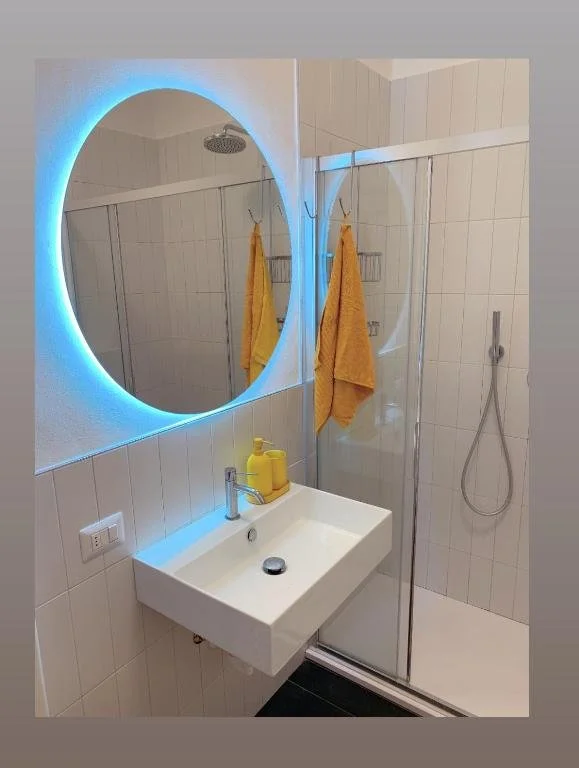 Bagno con grande specchio rotondo con luci LED blu, lavabo bianco con due dispenser di sapone gialli, due asciugamani gialli appesi, shower con cabina in vetro e doccia a parete, piastrelle bianche e pareti chiare.