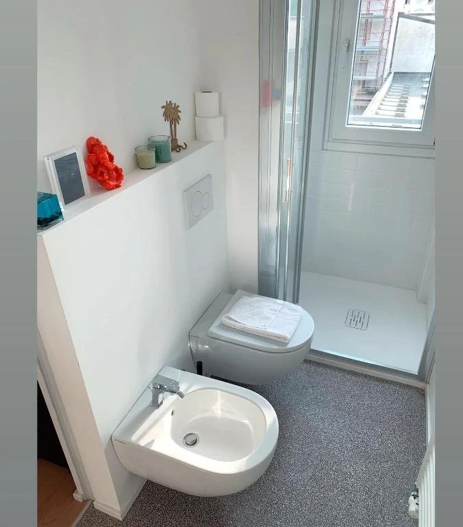 Bagno moderno con toilette e bidet, doccia con porta scorrevole, finestra, ripiano con decorazioni e asciugamani