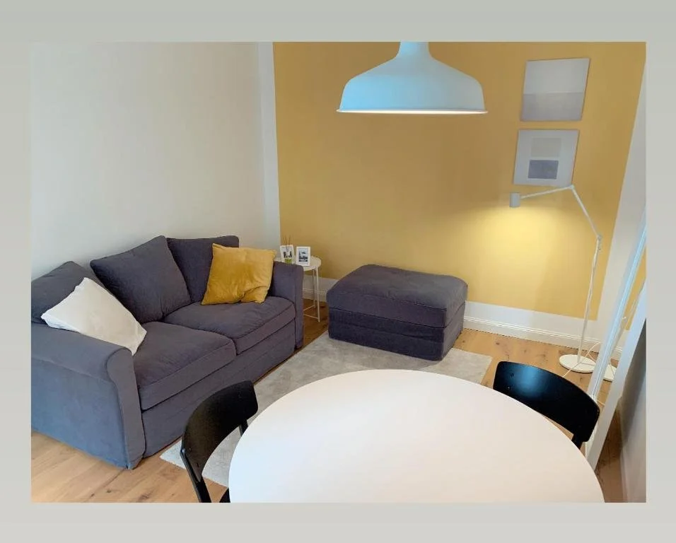 Soggiorno con divano grigio, pouf abbinato, lampada bianca da terra, tavolino con cornici e oggetti, parete gialla con quadri, tavolo rotondo bianco, sedie nere.