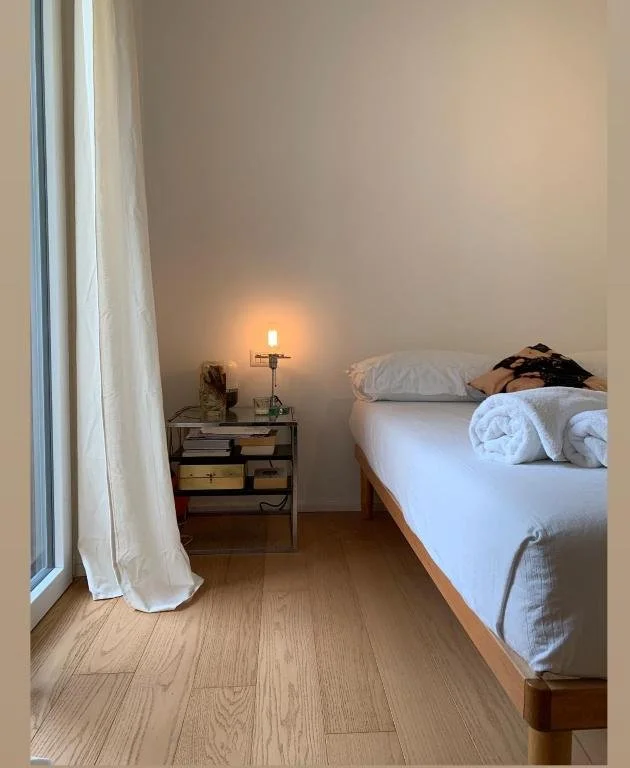 Camera da letto con letto matrimoniale, notte con lampada accesa, tavolino con libri e oggetti, finestra con tenda bianca, pavimento in legno.