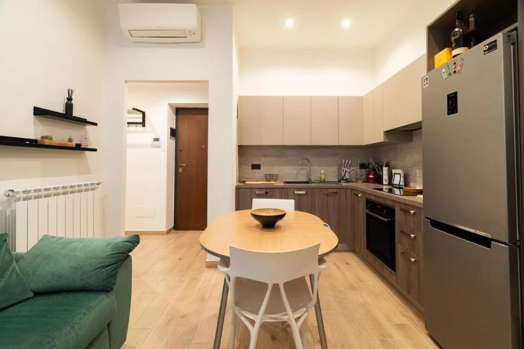 Cucina moderna con armadietti beige e castani, frigorifero in acciaio, tavolo rotondo con sedie bianche, divano verde, mensole nere con decorazioni, e pareti bianche con pavimento di legno