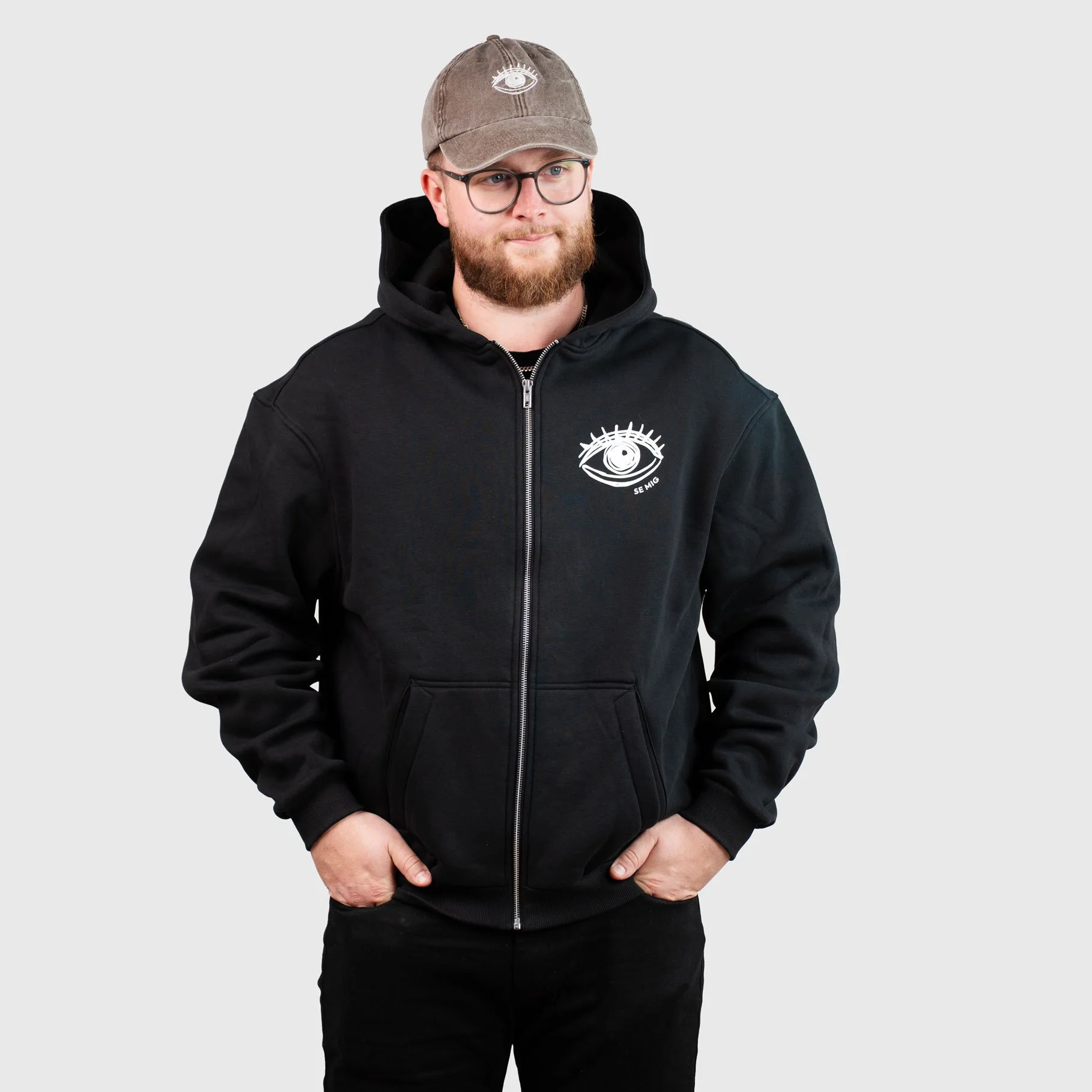 Man i keps och svart zip-hoodie med ögonlogga, framifrån