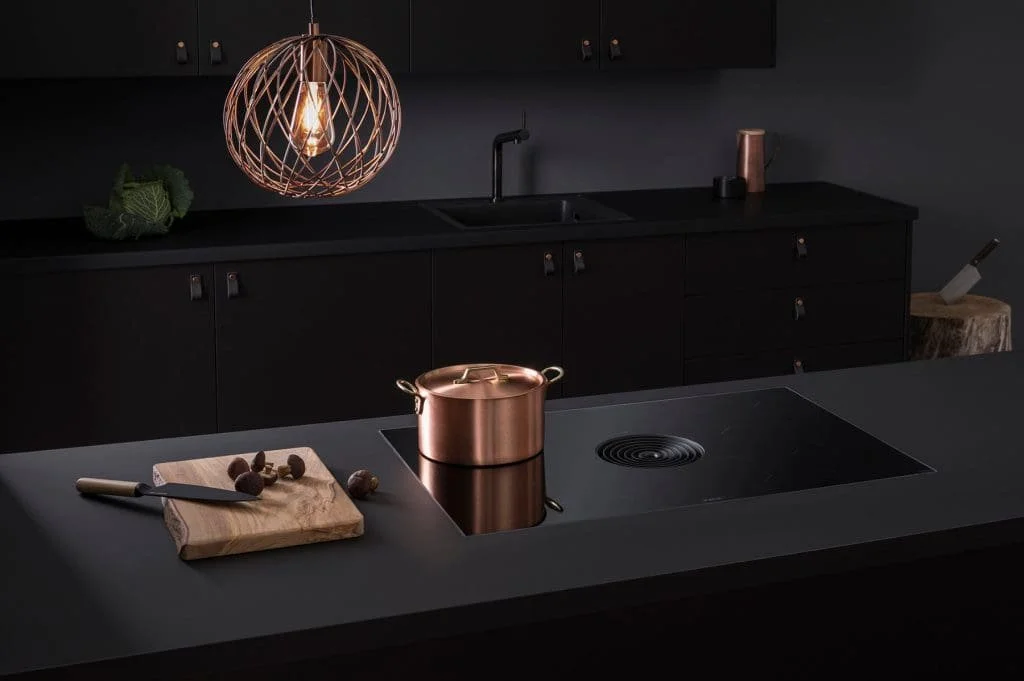 Modern black kitchen with copper cookware, wooden cutting board, and a decorative hanging light fixture.