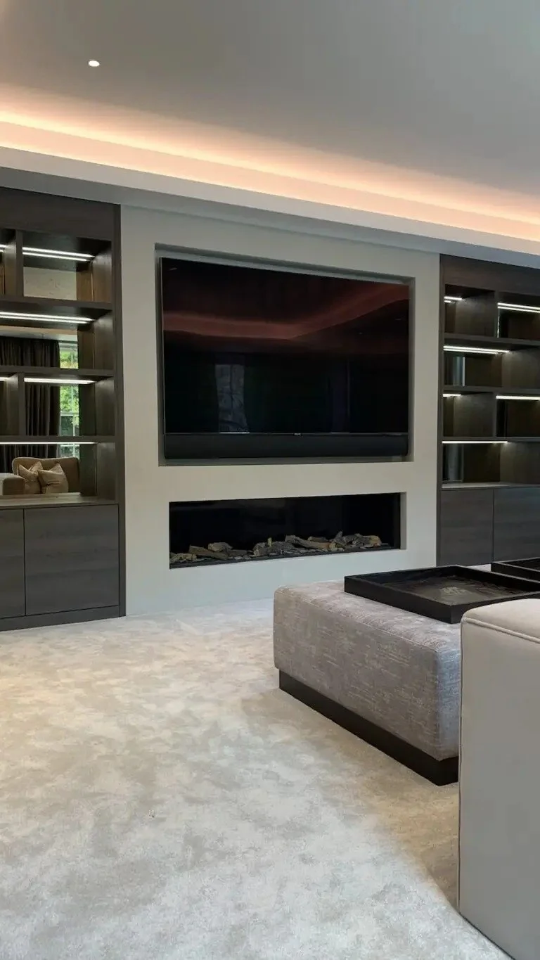 Photo of a modern living room with a large flat-screen TV mounted above a built-in fireplace with decorative stones. The room has a light-colored carpet, a beige ottoman with a tray on top, and dark-colored built-in shelves with lighting on either side of the TV.