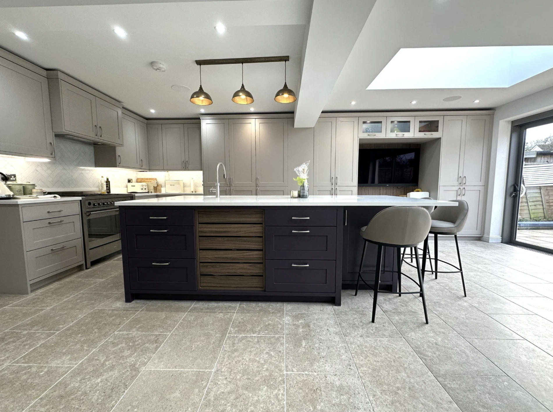 Modern kitchen with gray cabinets, a large island with a white countertop, a sink, and two bar stools. There are pendant lights hanging above the island and a skylight allowing natural light. The room features tiled flooring and a sliding glass door leading outdoors.