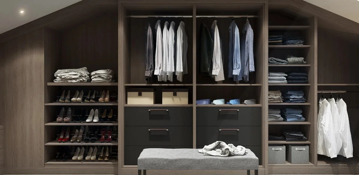 A large wooden closet with various clothing items and shoes organized on shelves and hanging rods. There are folded shirts, blouses, and pants, along with high heels, dress shoes, and storage boxes. A gray bench with a few clothes on top is in front of the closet.