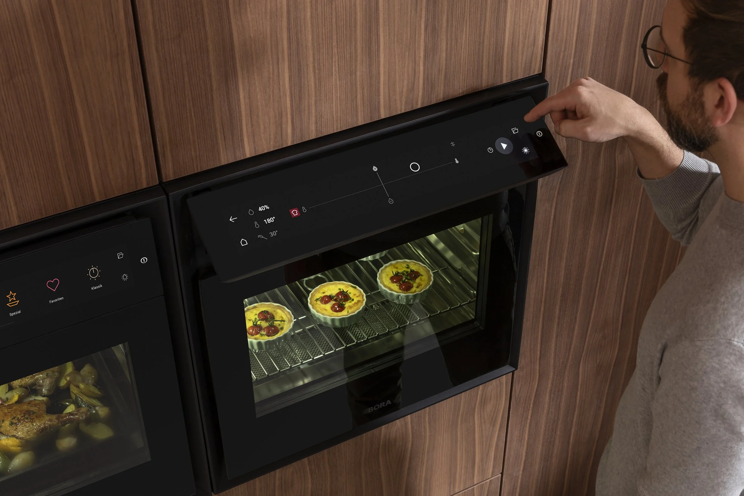 A man operating a modern black oven with a digital touchscreen display, baking three small vegetable quiches on a wire rack inside, in a wooden kitchen.