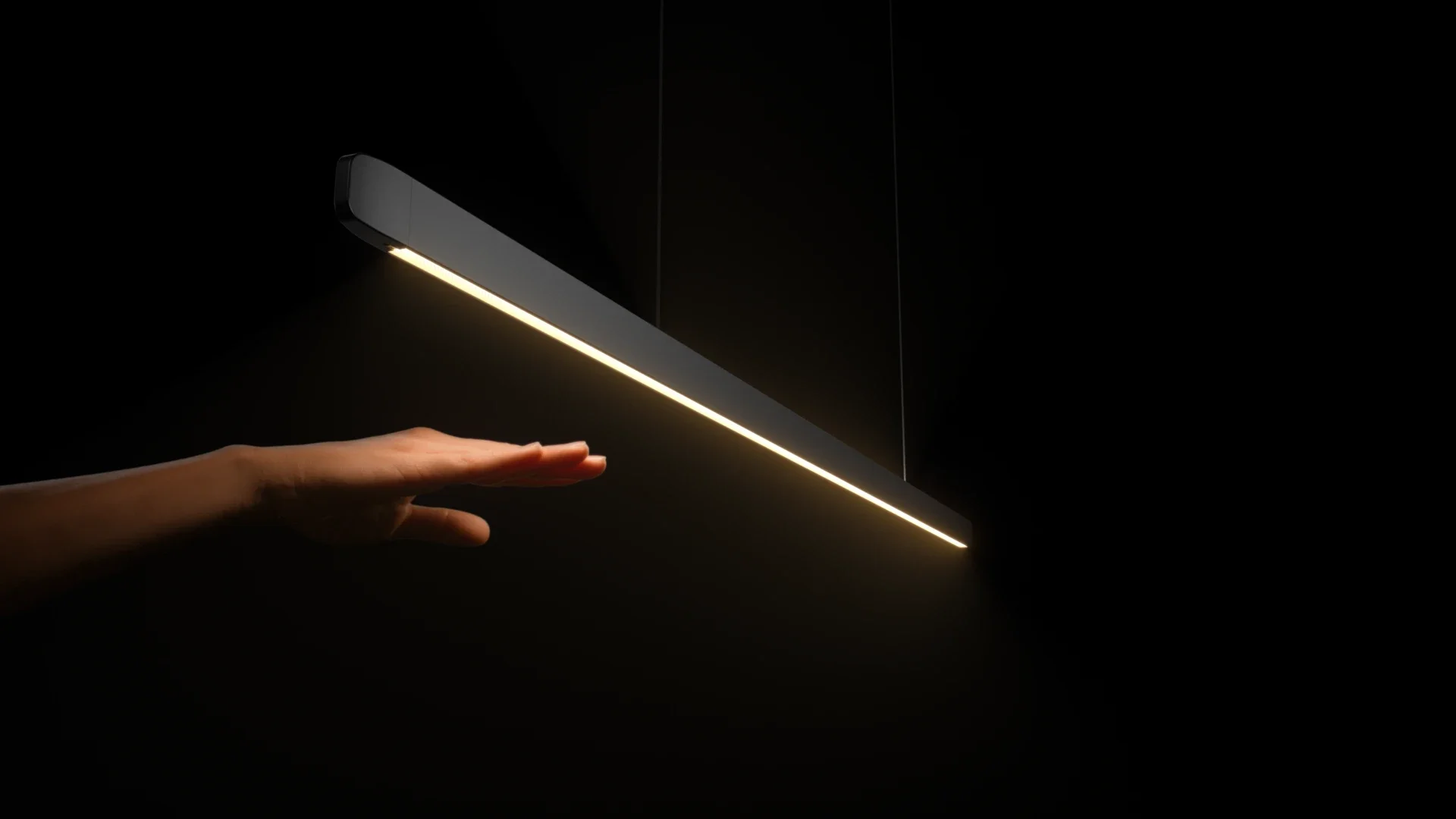 Person's hand reaching out towards a modern rectangular pendant light emitting a warm glow, against a dark background.
